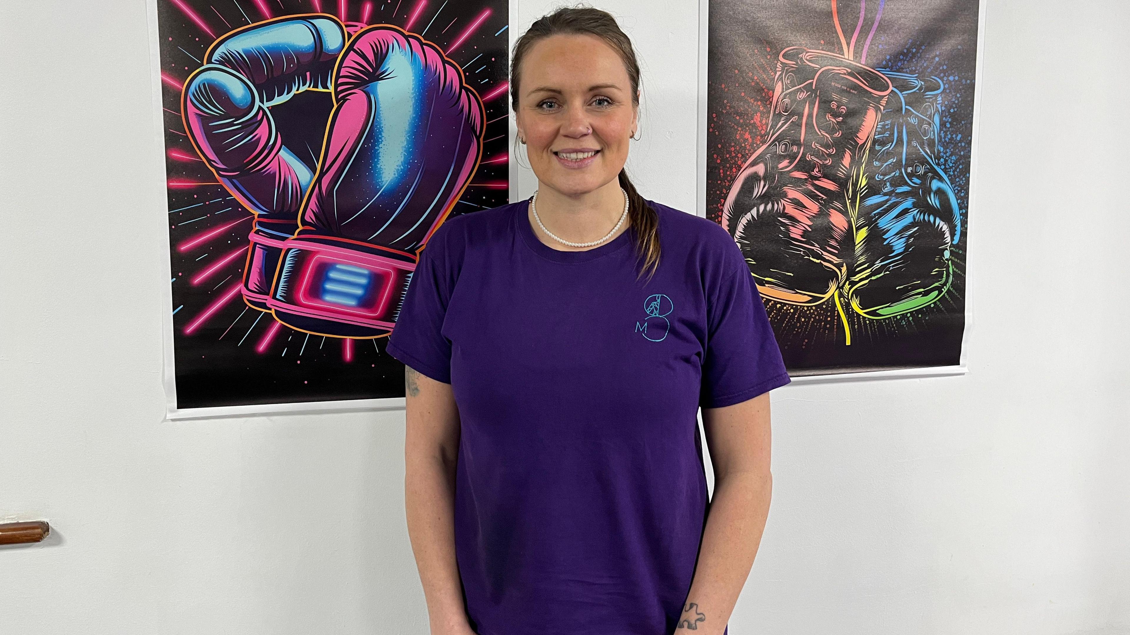 Michelle Minaides standing in front of boxing art work in a white room wearing a dark purple T-shirt smiling at the camera.