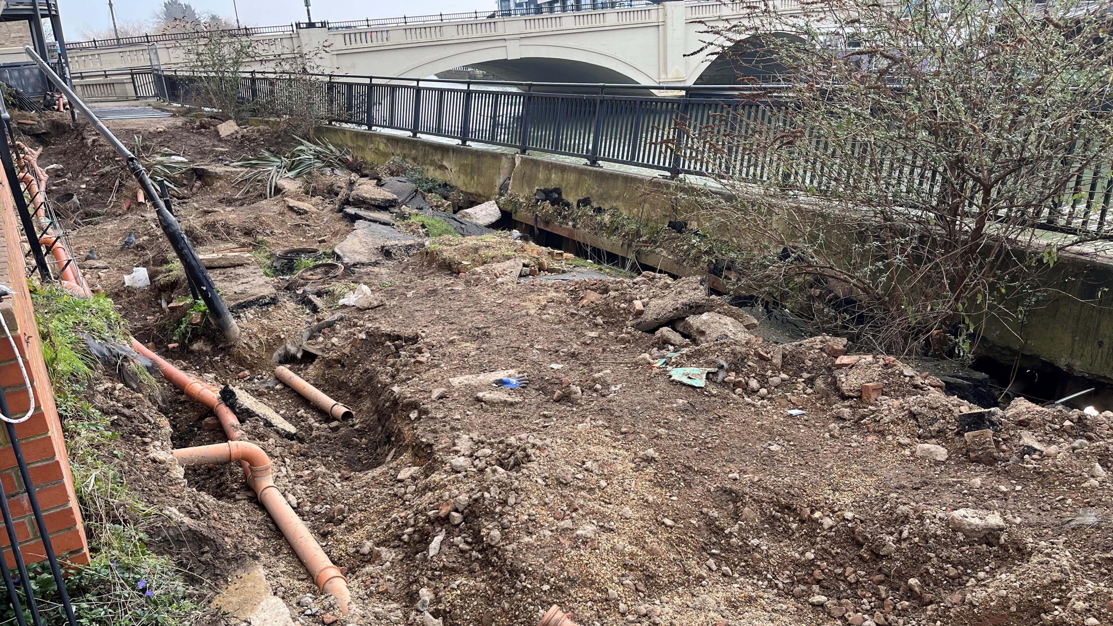 The front of the apartment block with a view from the other side of the flats, ground is earth that has been dug up to expose brown sewage pipes, a concrete wall and a black metal fence are badly bowed in the middle.