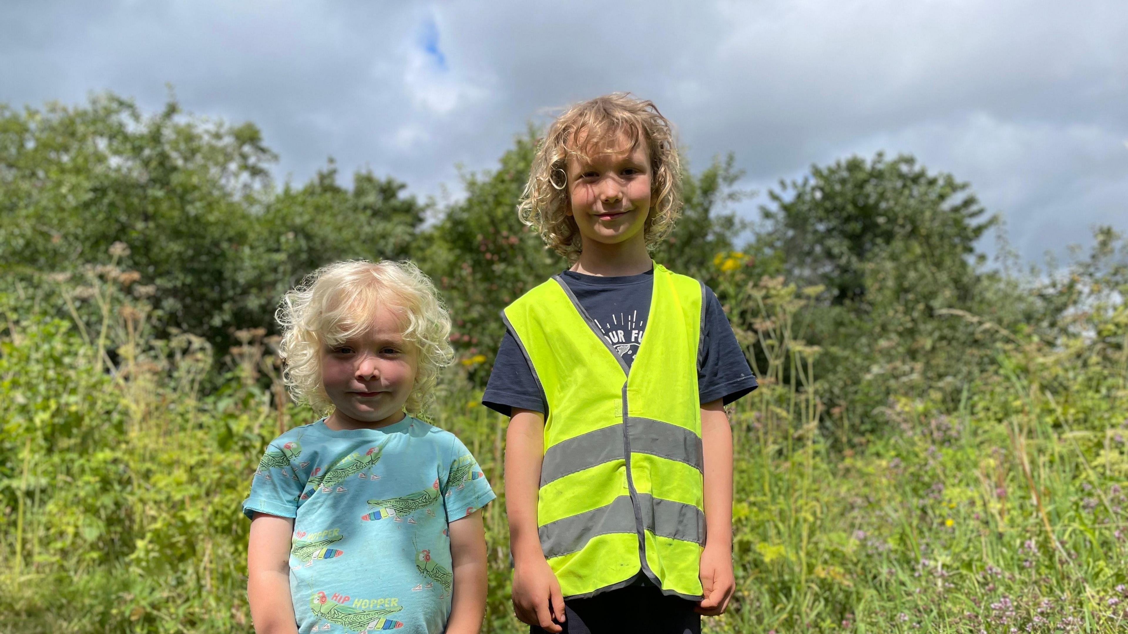 Two young boys, one in a pale blue T-shirt, the other wearing a hi-viz tabard, stand on rough ground looking at the camera.