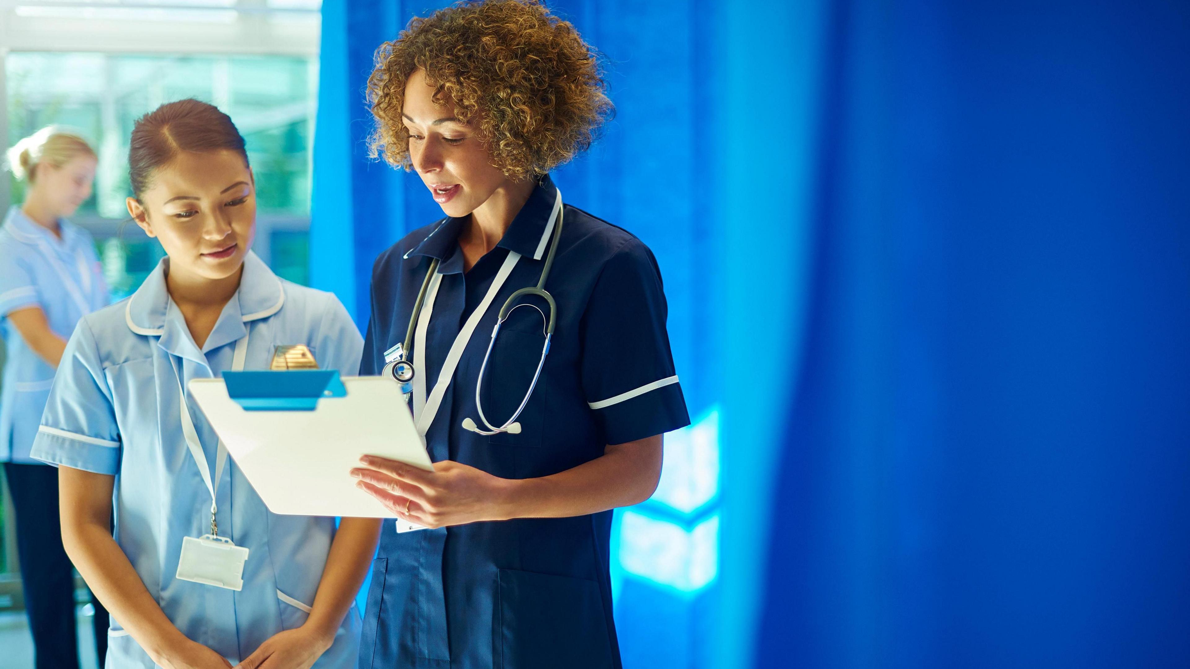 A nursing sister supervises a young ward nurse standing at the foot of a hospital bed, chatting about the medical chart that she is holding.