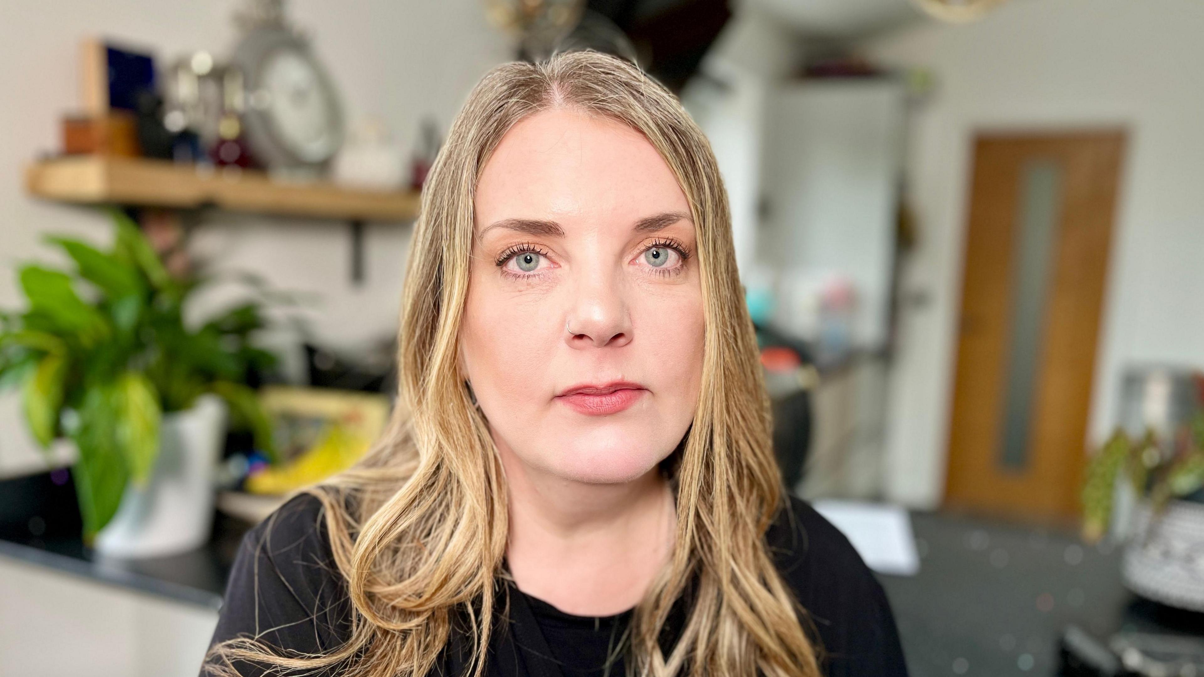 Louise Longster, who has blue eyes and blonde hair, looks direct at the camera. She is wearing a black top. She is sitting in a room and a pot plant, shelf and door are visible behind her.