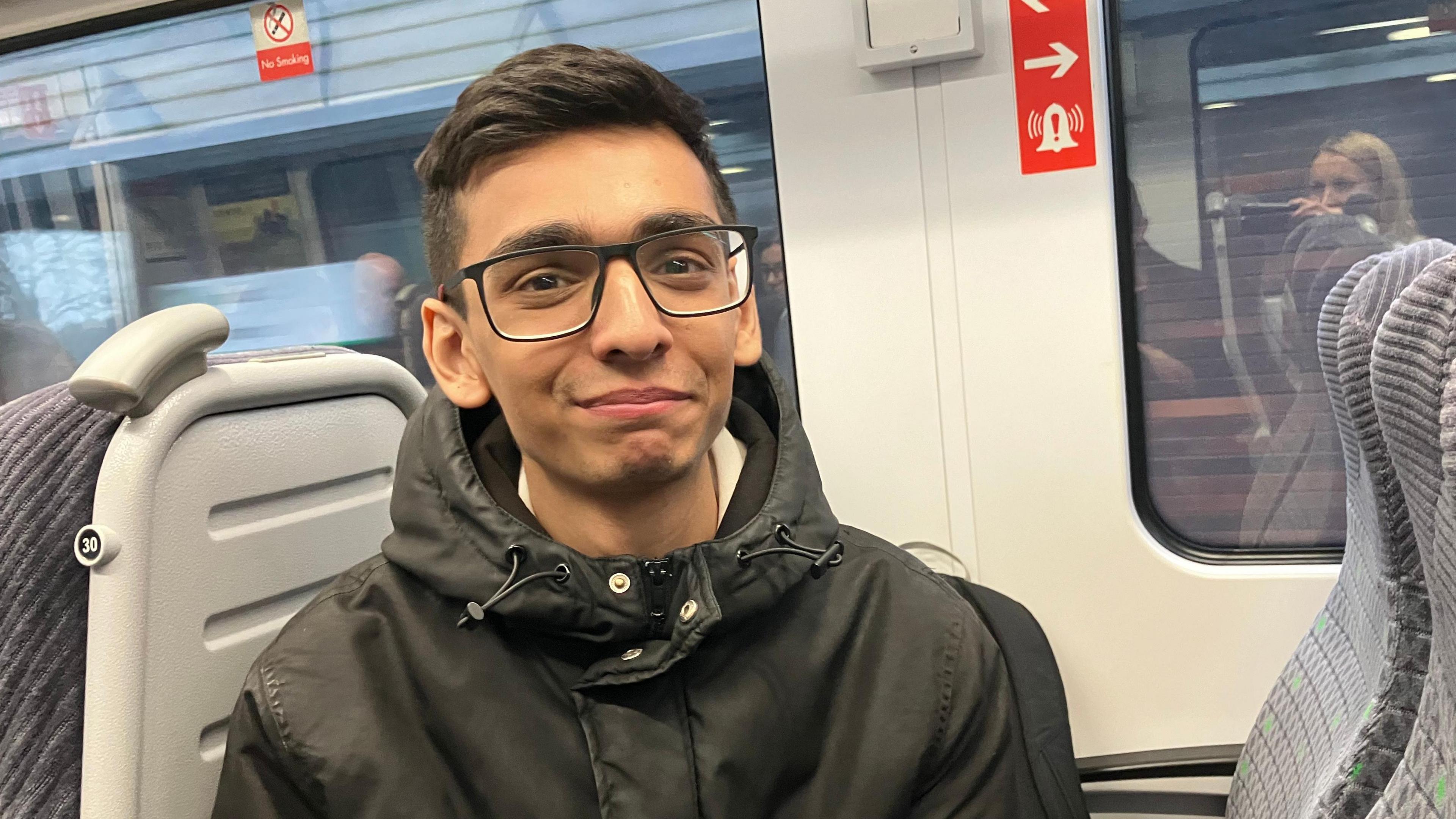 Eesa Ashraf with short dark hair and wearing glasses, a dark hooded jacket done up to his neck. He is sitting in a railway carriage 