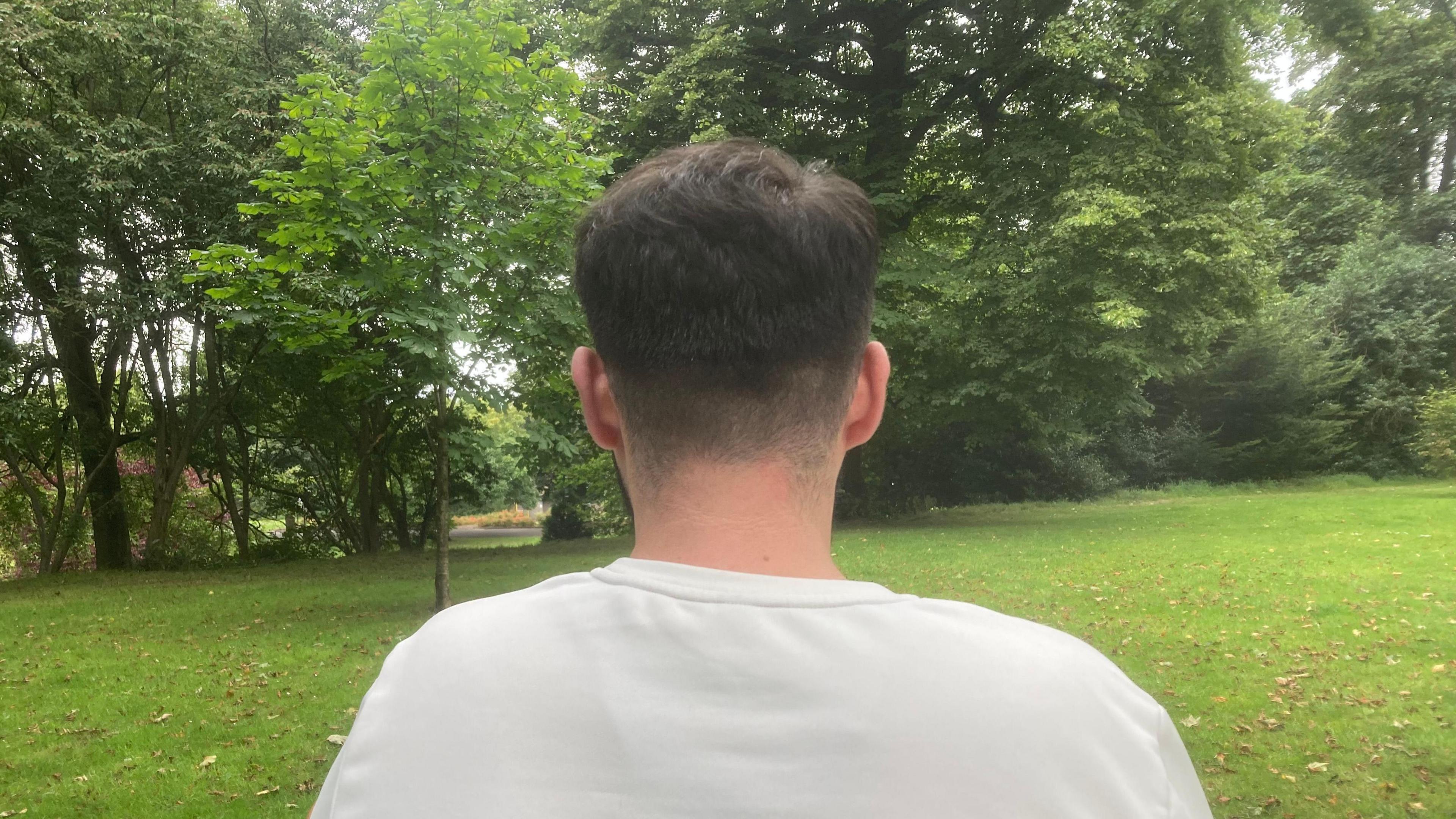 A man standing in front of some trees. He is turned away from the camera so none of his face is visible, and he is wearing a white shirt. 