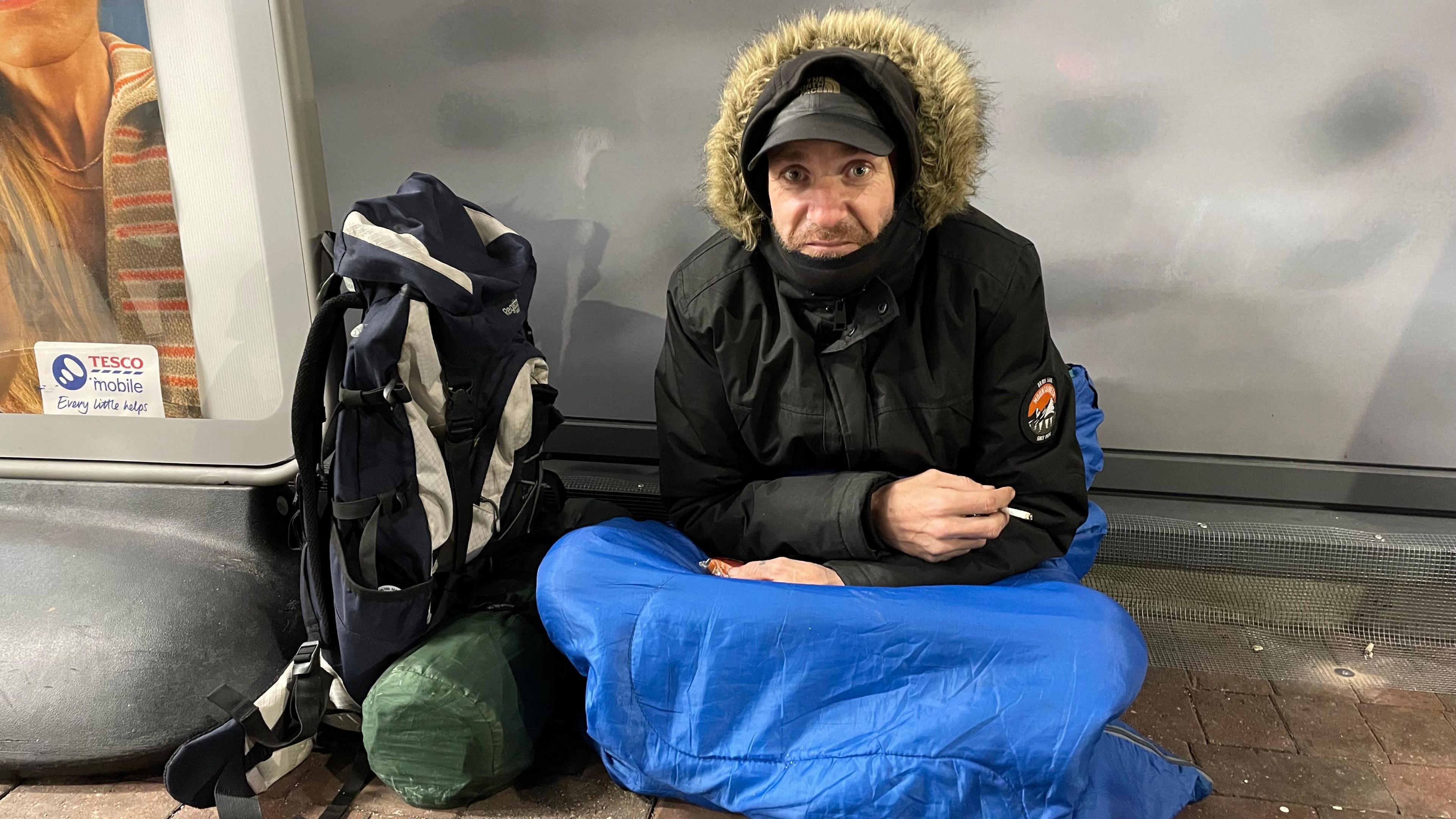 Rich is sitting outside a supermarket in a sleeping bag and warm coat