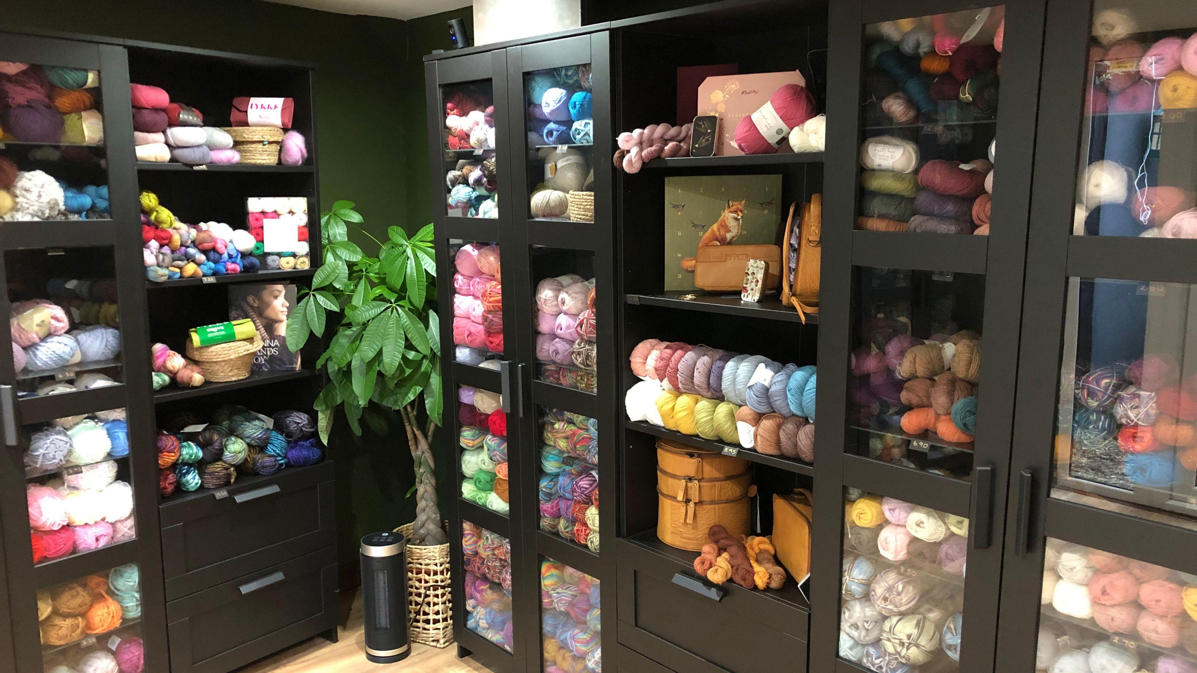 A picture of the shop's shelves. The shelves are black and are holding tens of yarn balls in all different colours. An indoor plant can be seen in the corner of the room between two of the shelves. Other shop items can be seen on the shelves. 