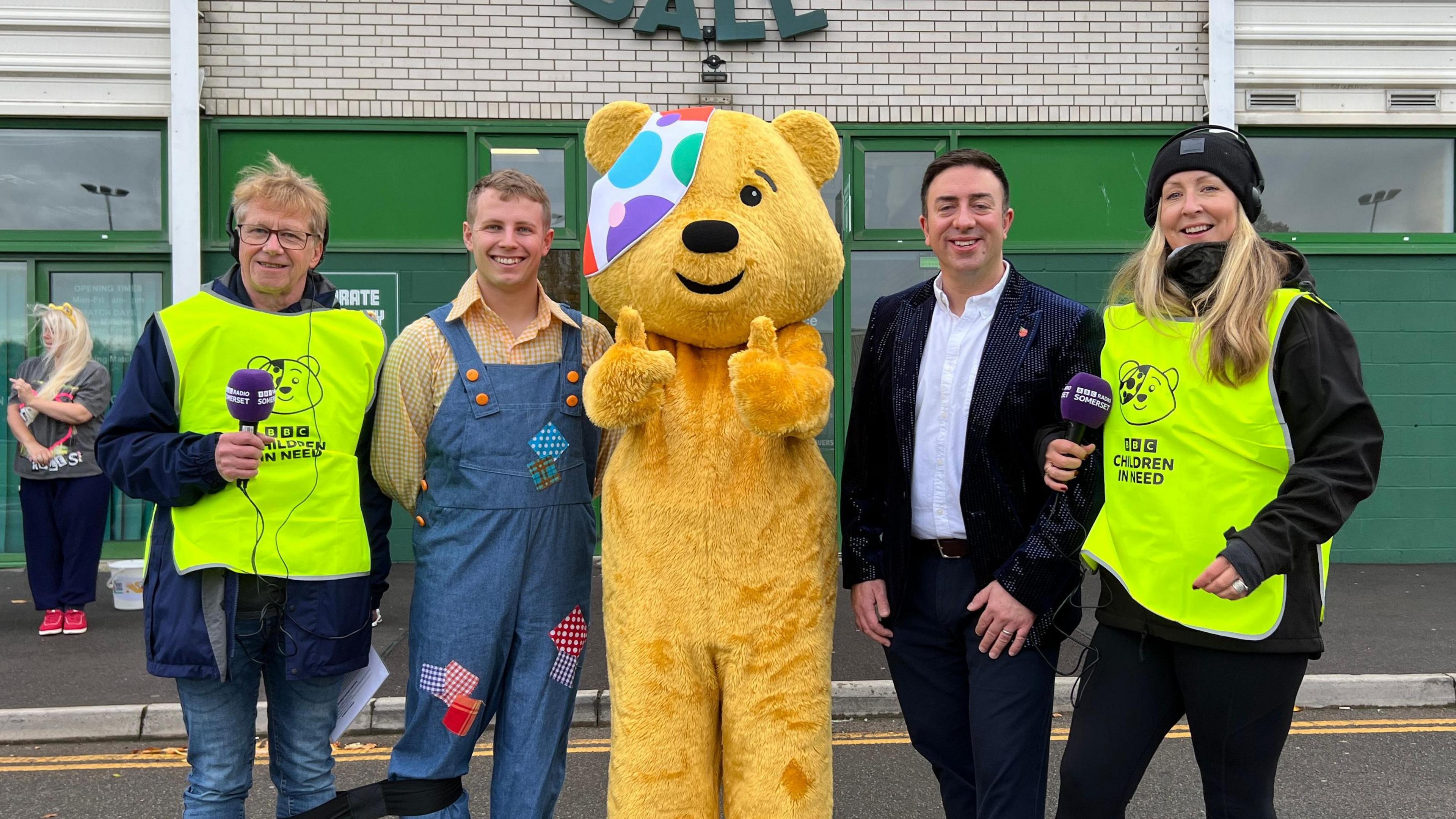 Four people are standing next to Pudsey bear at Huish Park.