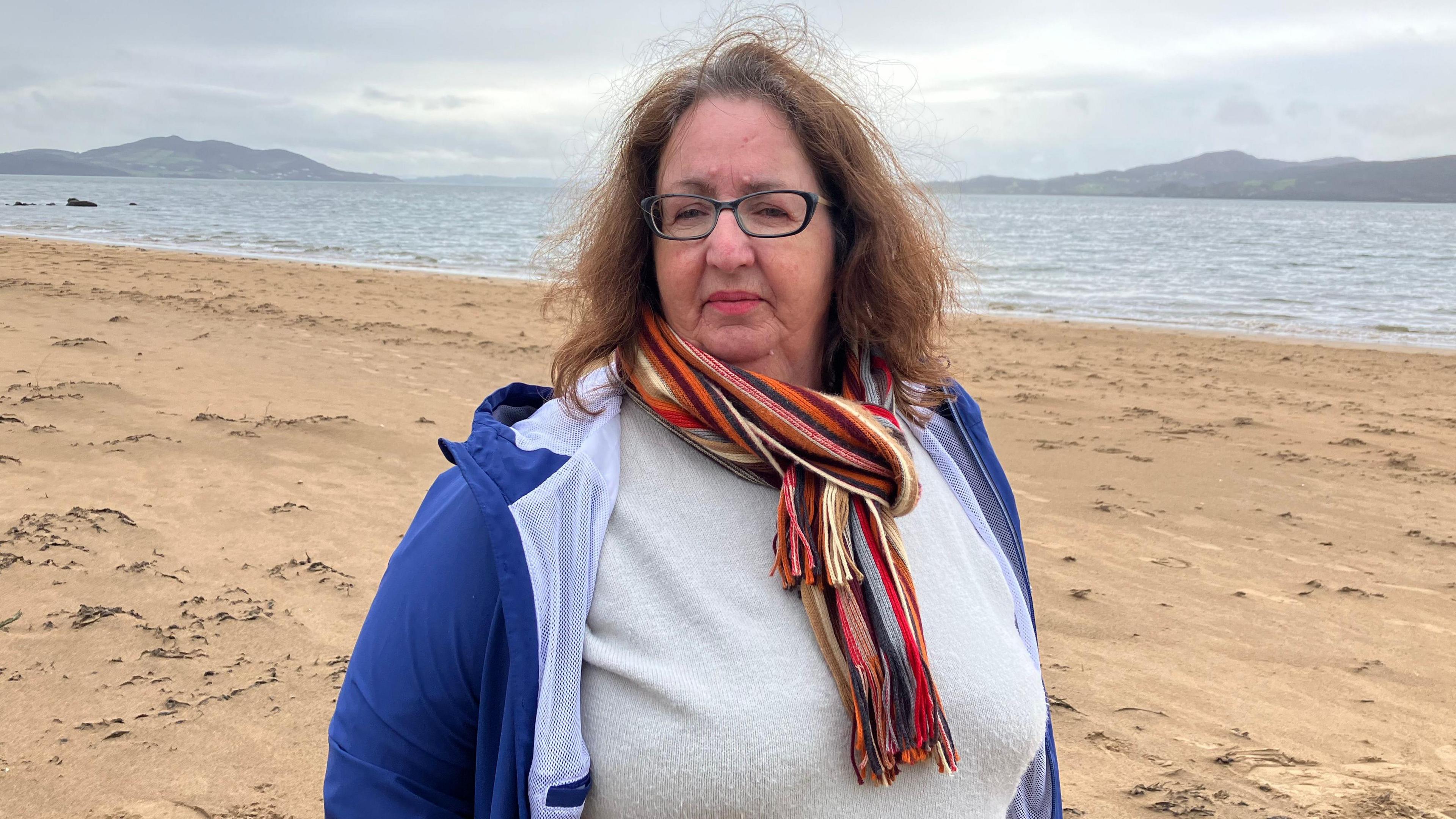 Andrea brannigan stands on a beach in Donegal, Ireland, she is wearing glasses and has long brown hair. Lough Swilly is behin her and the sands of the beach are also seen. In the distance is the donegal coastline. She is wearin a blue coat, grey sweather and multi coloured scarf.