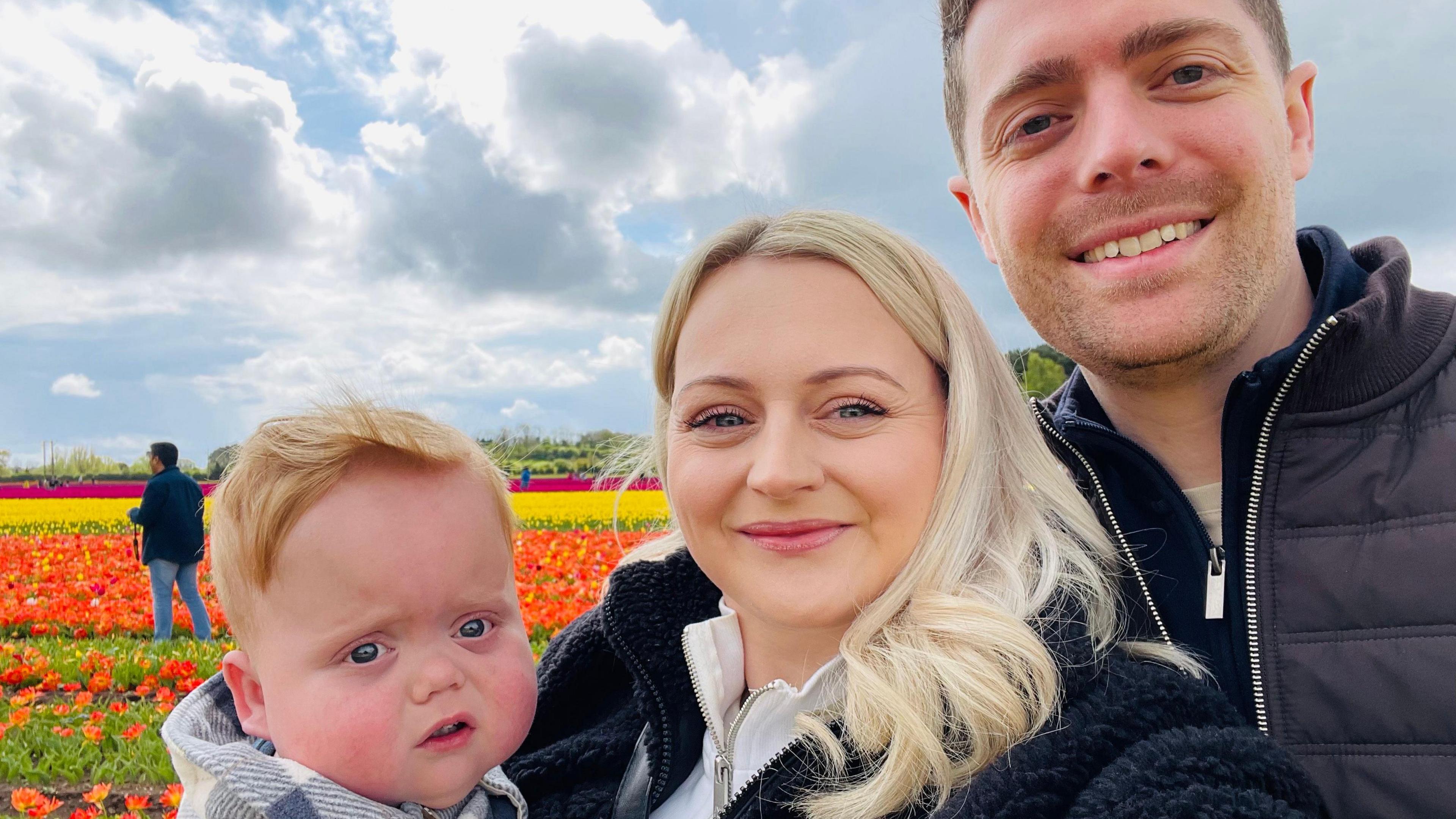 Reece and Shona are smiling at the camera while holding Ari. Behind them is a field of vibrant red, orange, yellow and pink flowers. Shona is wearing fluffy black jacket and Reece wears a black zip up coat.