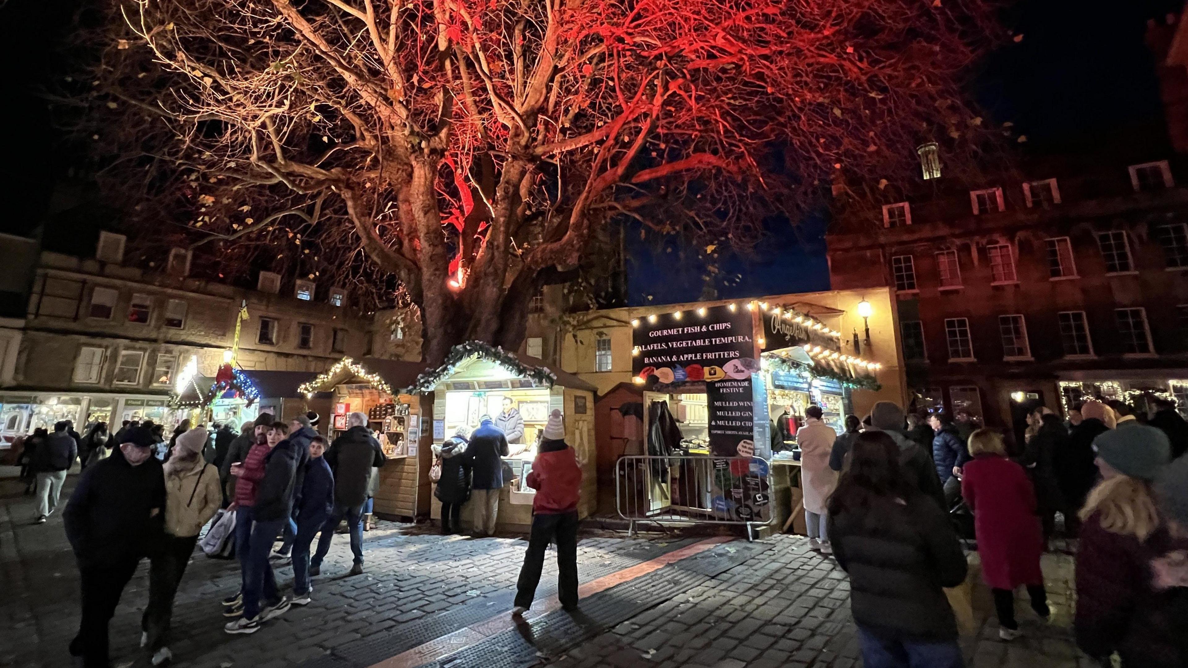 Dozens of people walk between stalls at the Bath Christmas Market. There are trees illuminated from below with festive lights and the stalls have tinsel and lights on their frontage