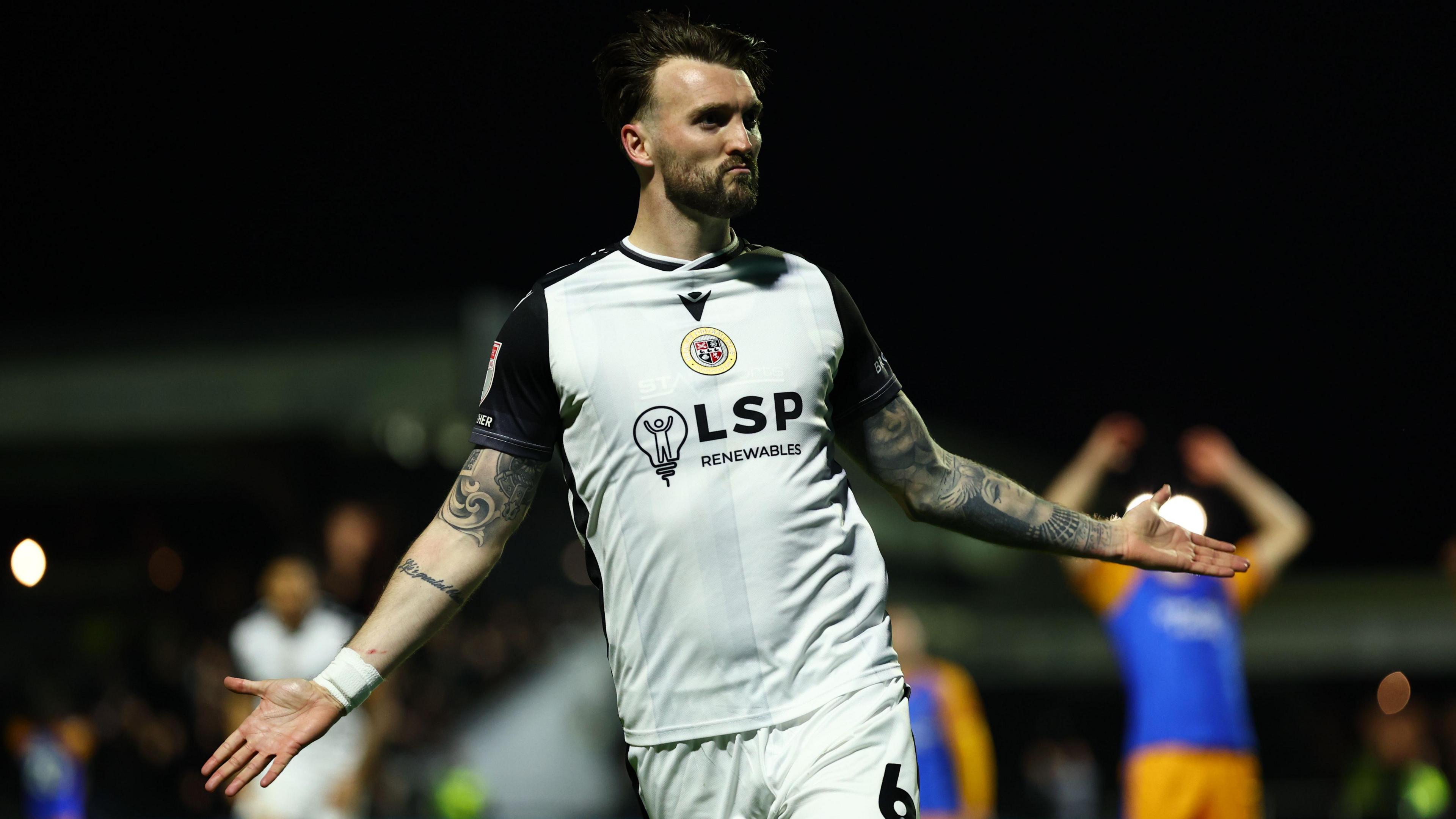 Kyle Cameron looks proudly towards supporters with arms outstretched in celebration, wearing home kit of white with black shoulders