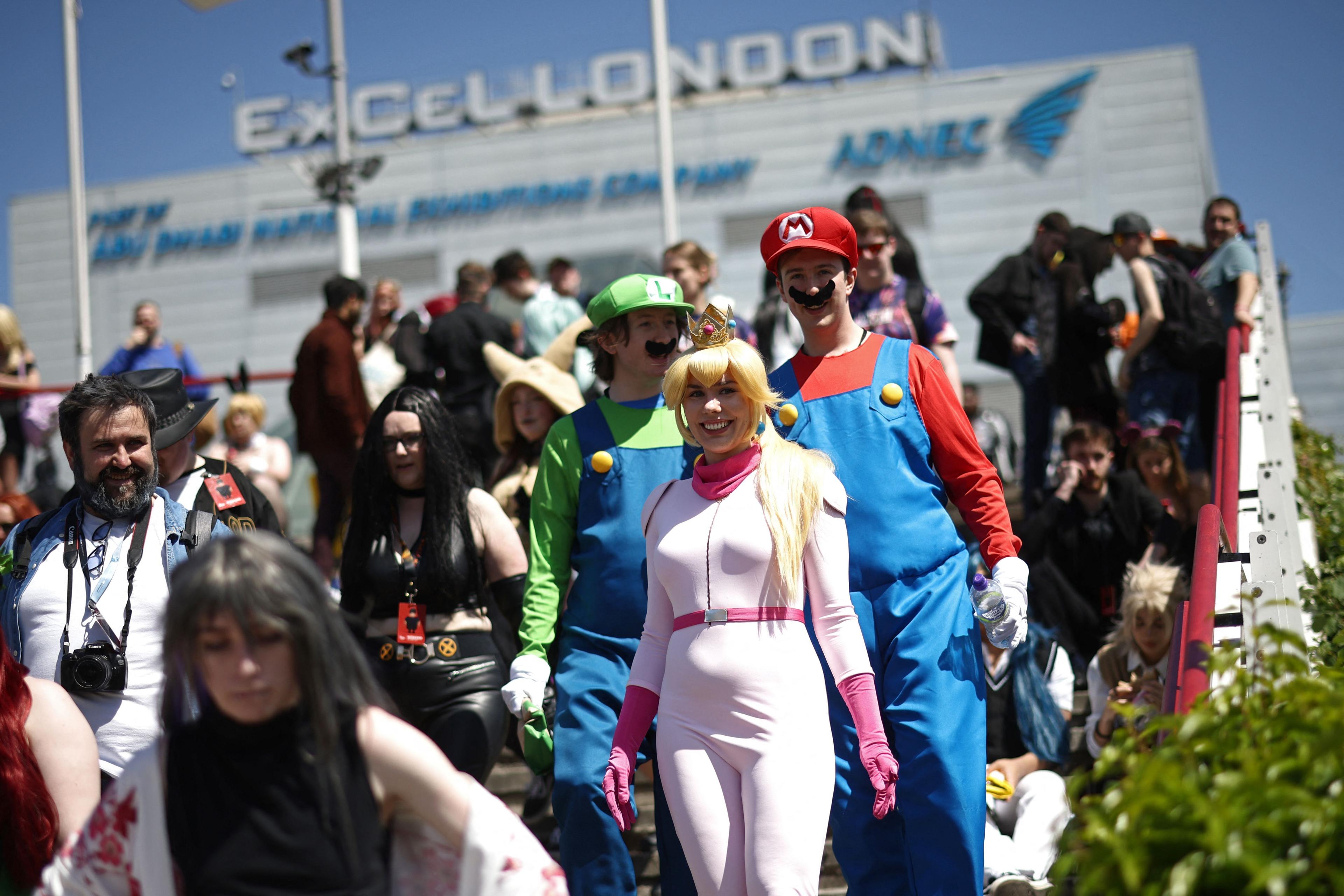 Outside of ExCel in London where Comic Con is held. The picture features people attending the convention, dressed up as various characters. The main people in the photo are seen dressed as characters from Super Mario Bros. A woman is dressed as Princess Peach, wearing a blonde wig, a gold crown and a light pink jumpsuit with dark pink gloves. Two men behind are dressed as Mario (right) and Luigi (left). Both men are wearing blue overalls and dark brown moustaches. Luigi is wearing a breen shirt underneath his overalls and a green hat with the letter "L" on it. Mario is wearing a red shirt under his overalls and a red hat with the letter "M" on it. Other people in various costumes surround them.