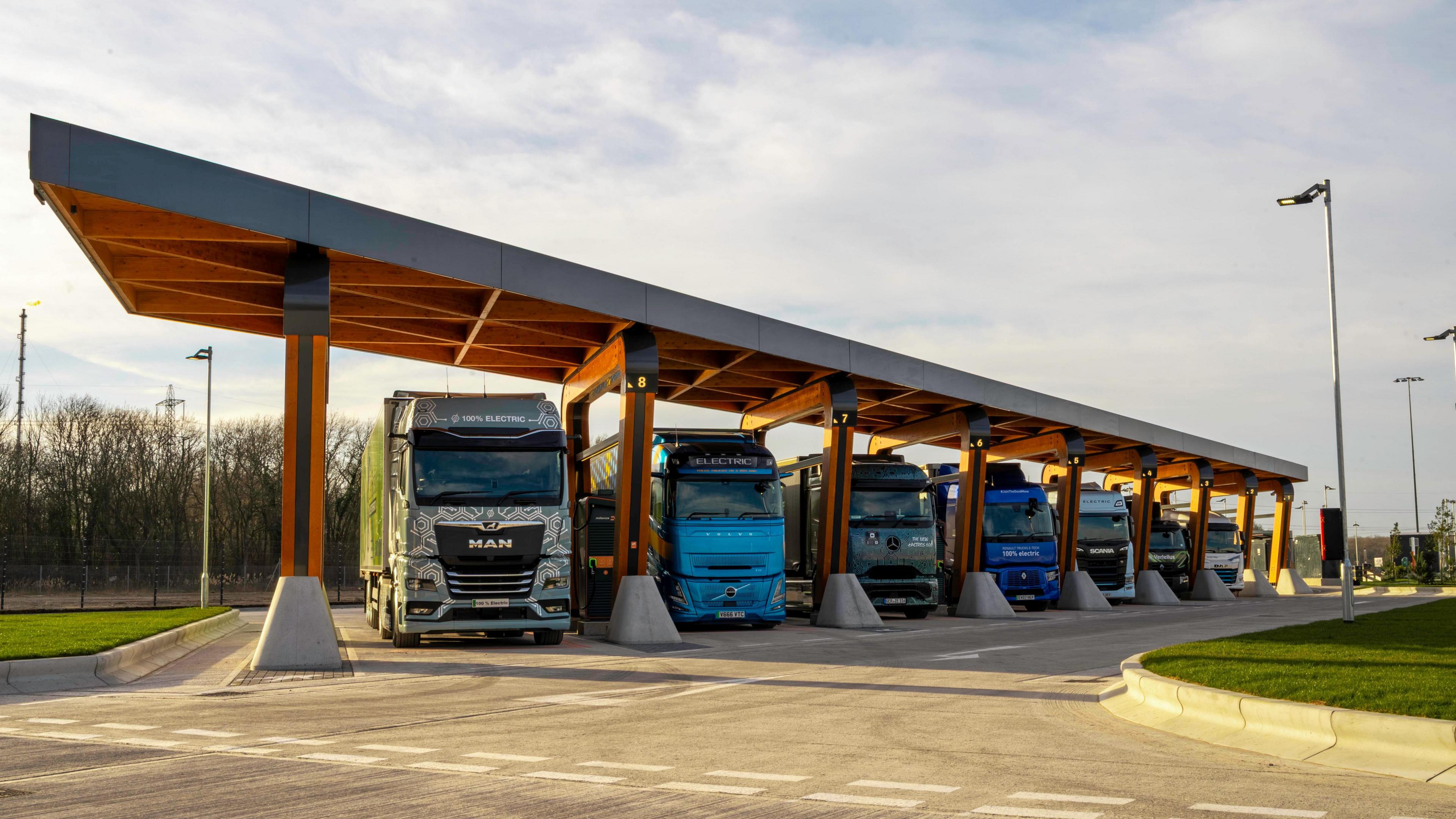 Seven HGVs lined up in seven electric charging points on industrial land. The design is orange and has a roof going over each port.