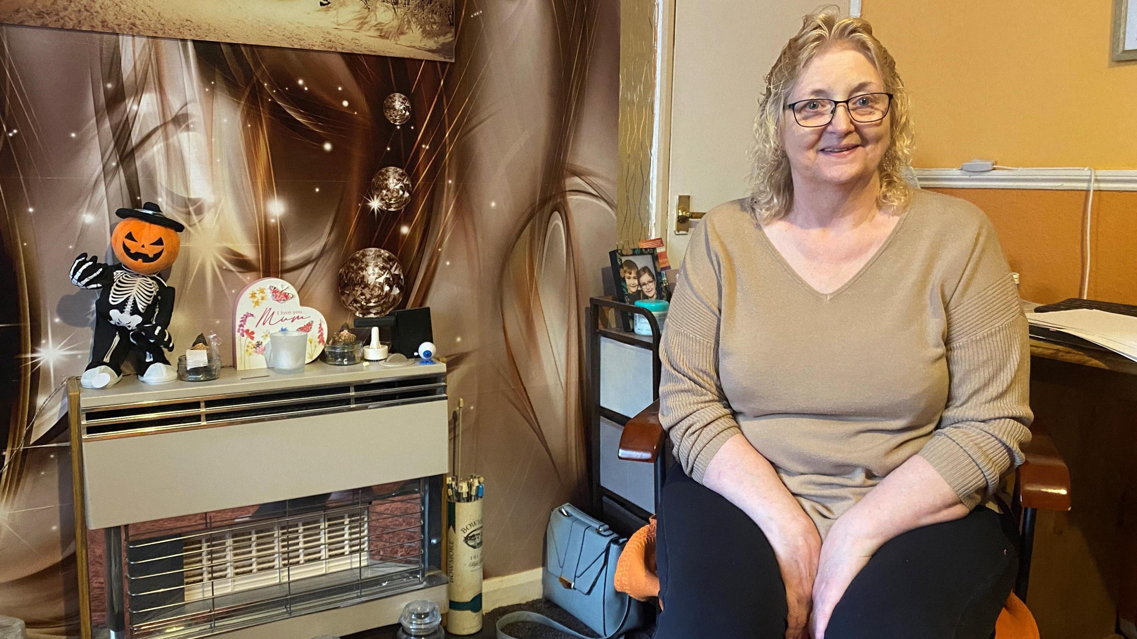 A woman in her sixties sit to the right of a gas fire. She's wearing a brown top and black trousers and is smiling at the camera 