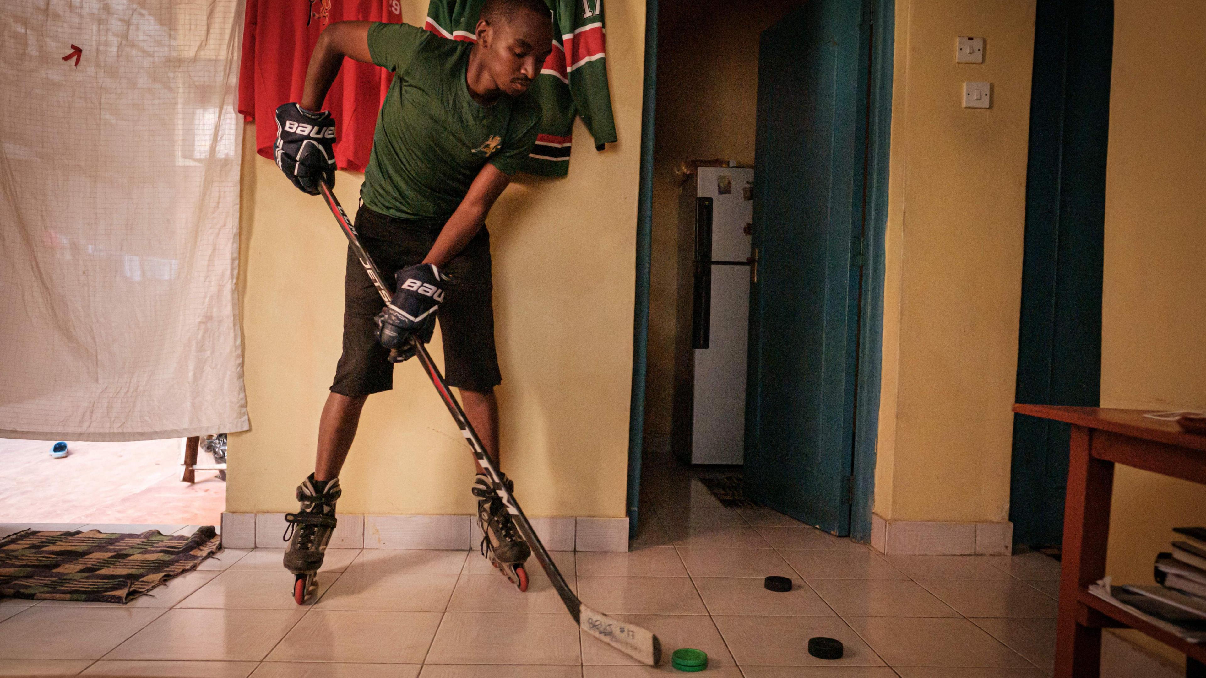 Benjamin Mburu, wearing a green T-shirt, black shorts, rollerblades and black ice hockey gloves, holds a stick and tries to control a puck on a tiled floor in his house