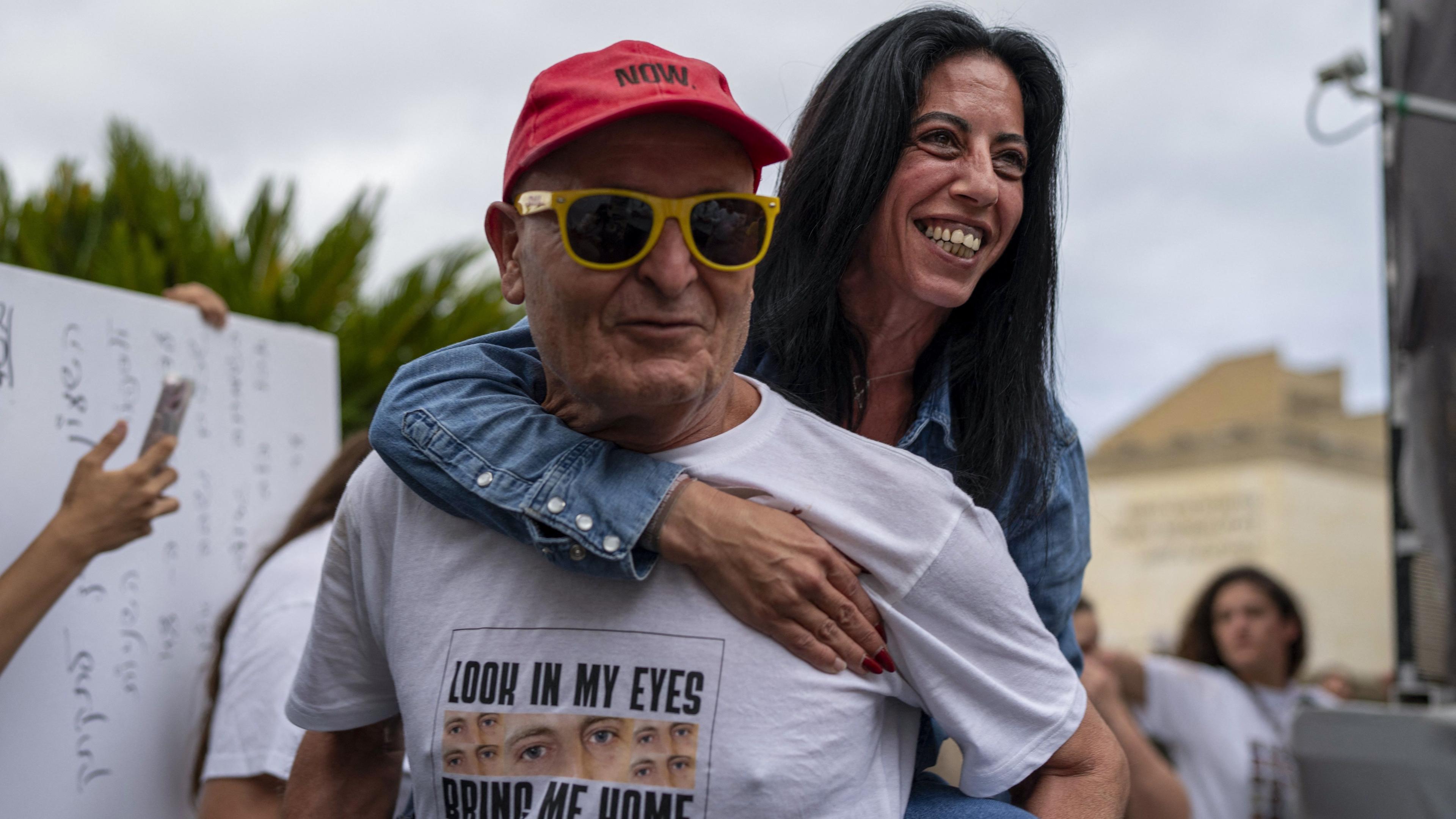 A woman jumps on the back of a man while smiling. The man wears a white t-shirt, red cap and yellow sunglasses.
