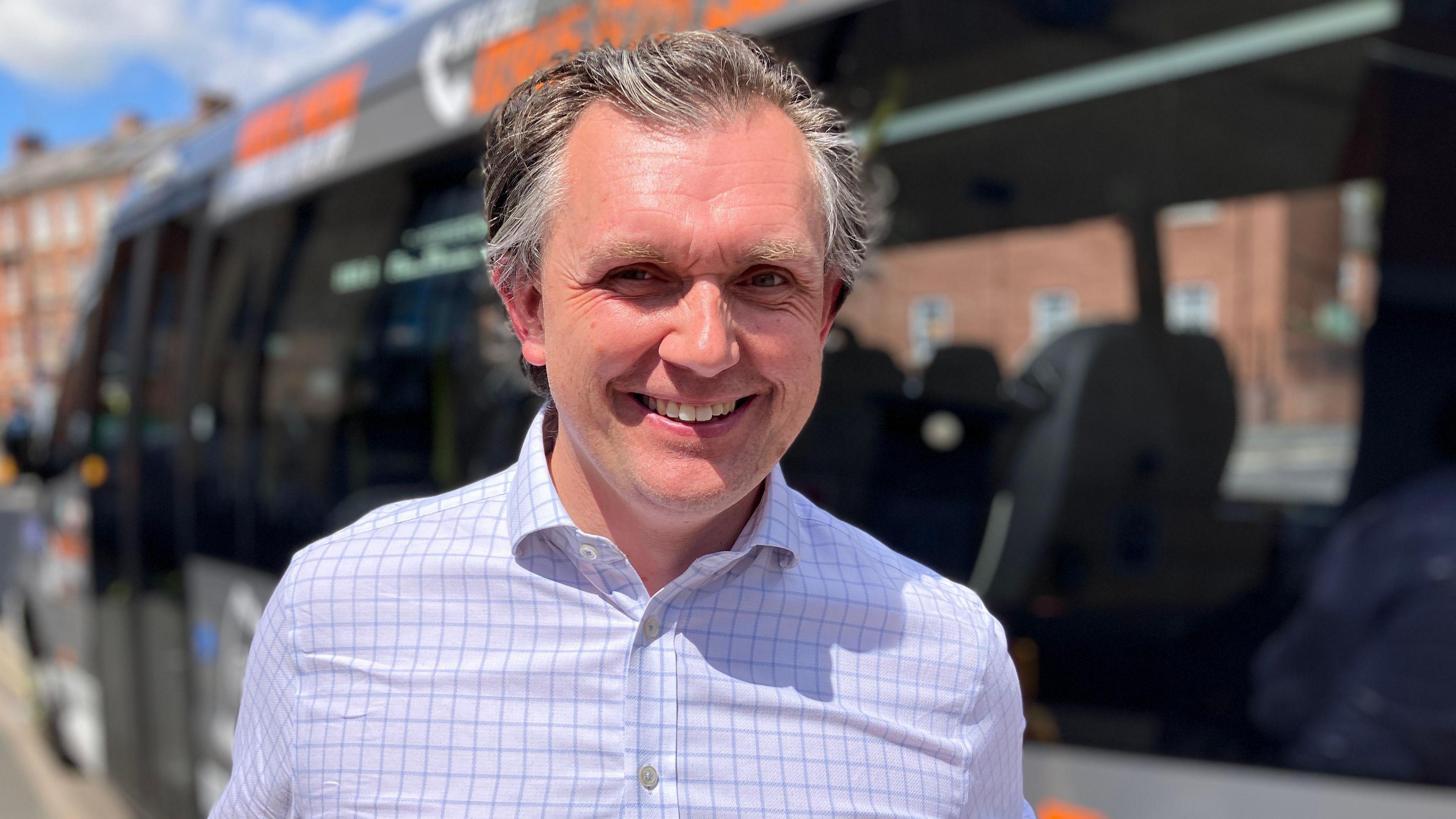 A man combed back grey hair and wearing a white and purple chequered shirt is smiling at the camera in front of a blurred bus behind him.
