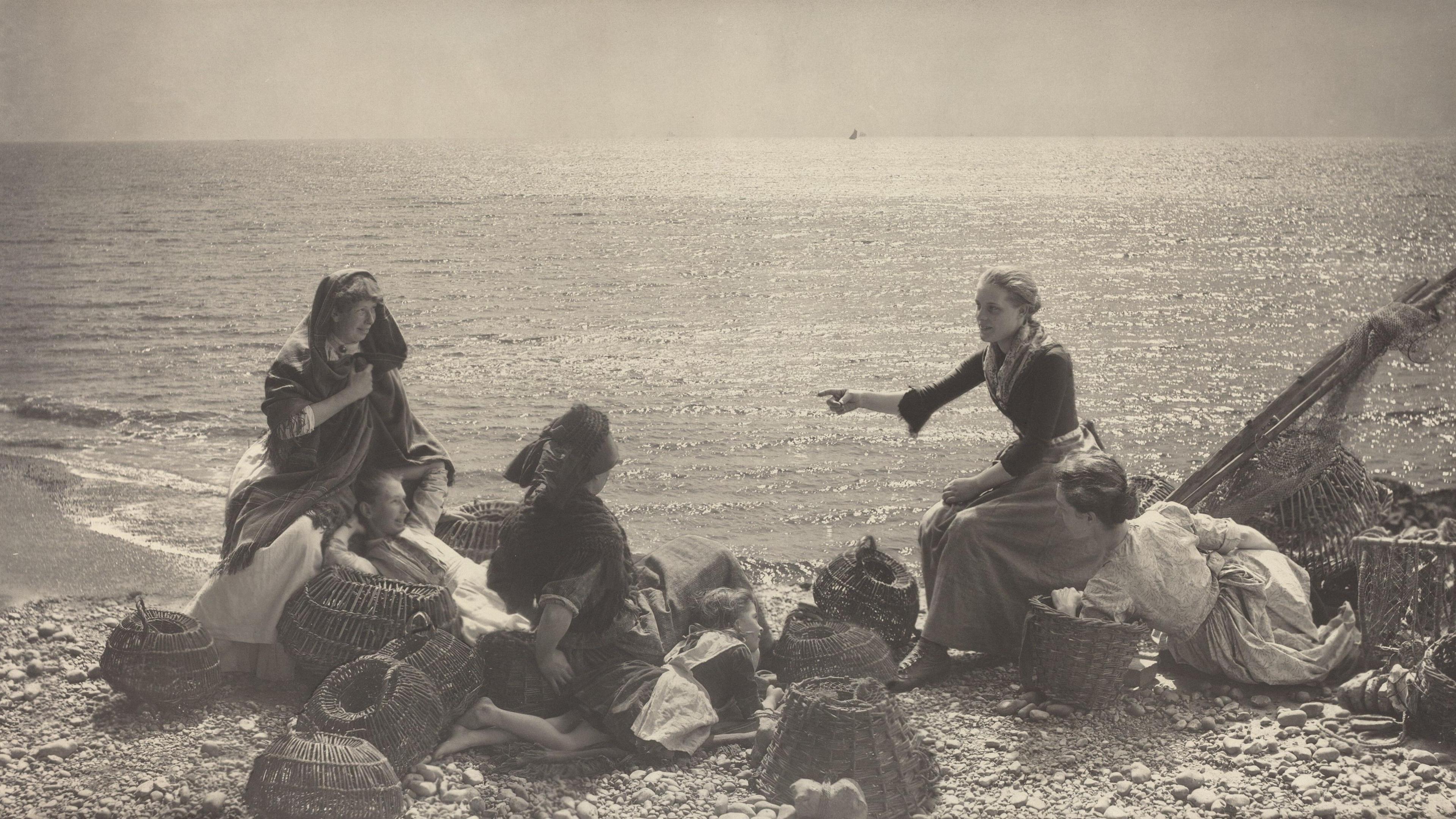 A sepia coloured photograph showing a group of women and children on a stony beach surrounded by baskets. They wear long skirts and dark tops, two of them with hats on.