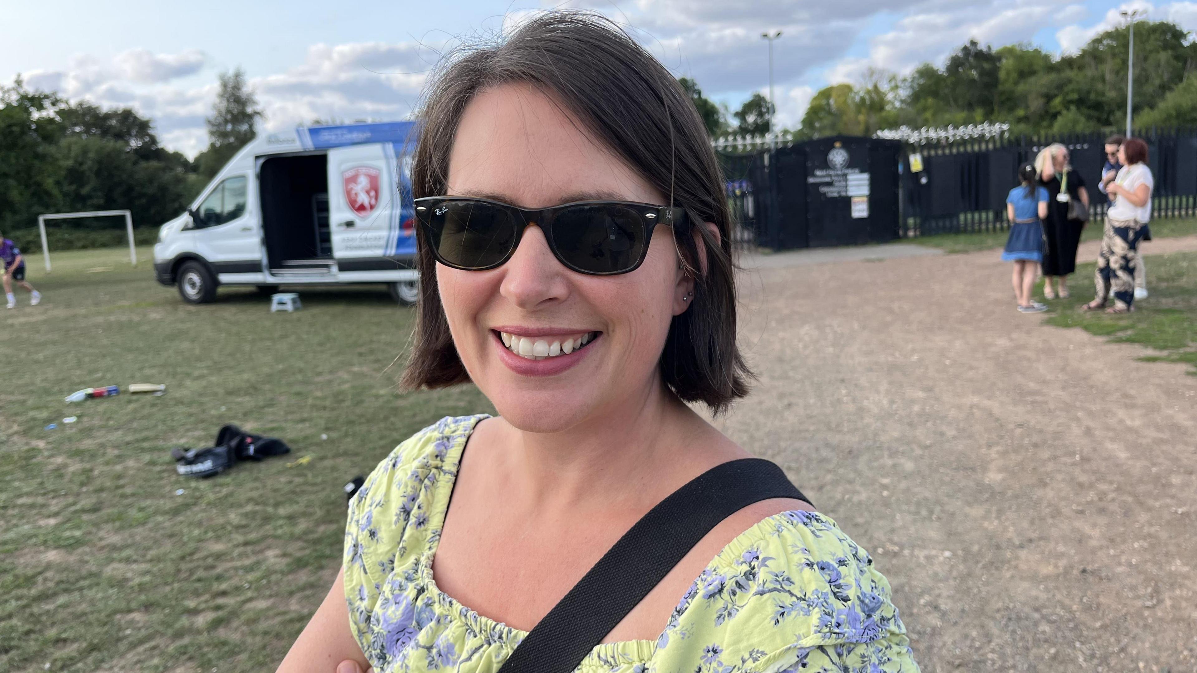 Jennie Fielder smiles into the camera. She is wearing sunglasses and a yellow and blue dress. She has short black hair.
