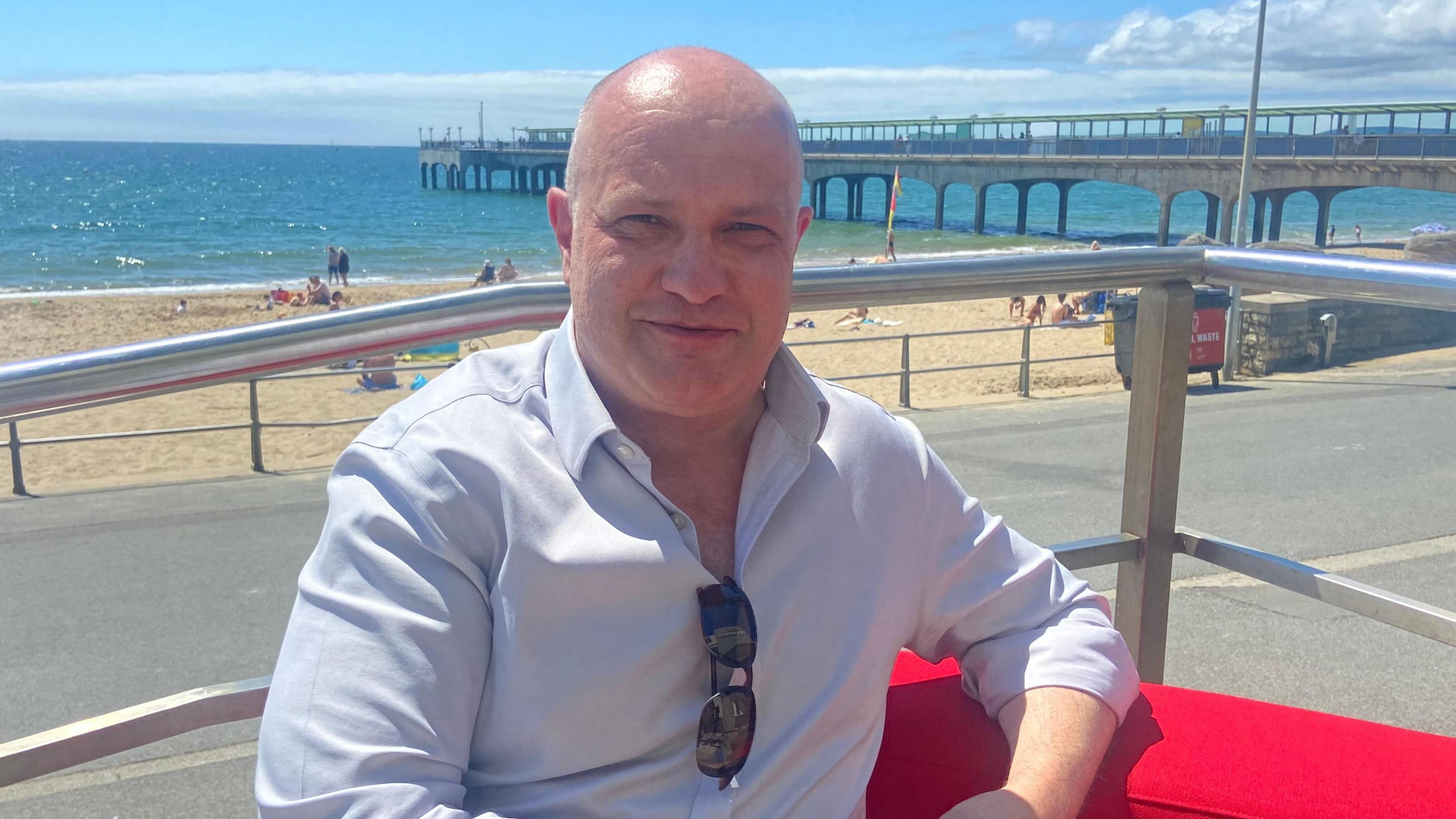 Martin Houlden wears a light blue shirt with sunglasses tucked into the front while sitting on a red sofa