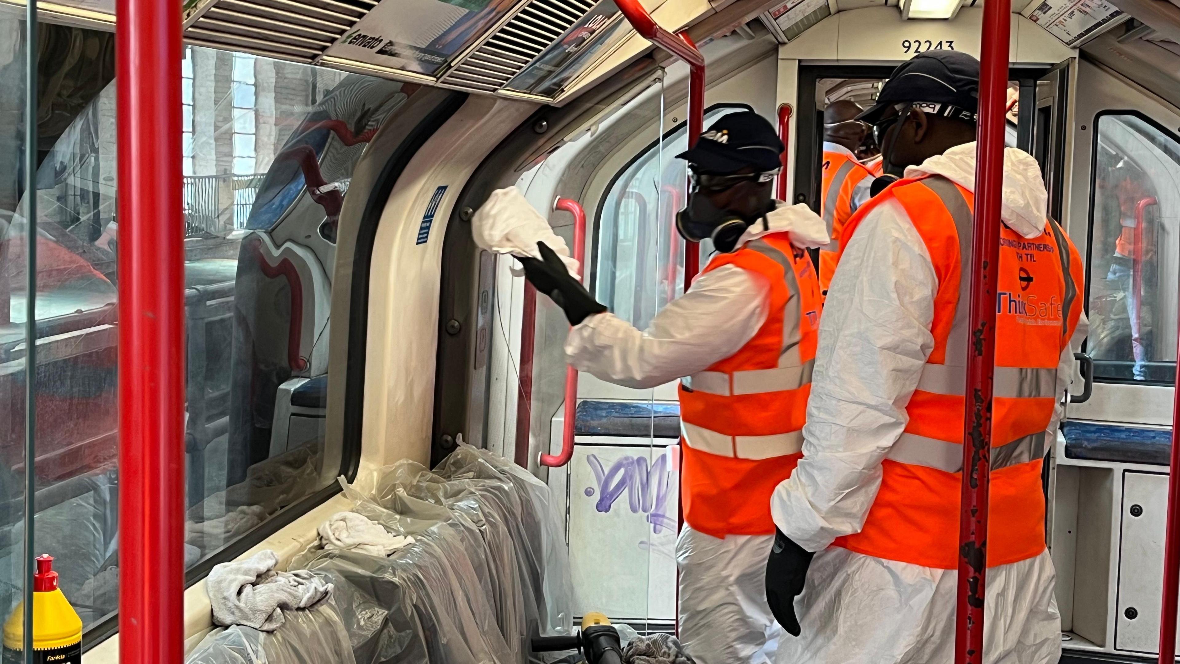 Two men dressed in white protective overalls and hi viz vests clean the graffiti inside a train with buffers