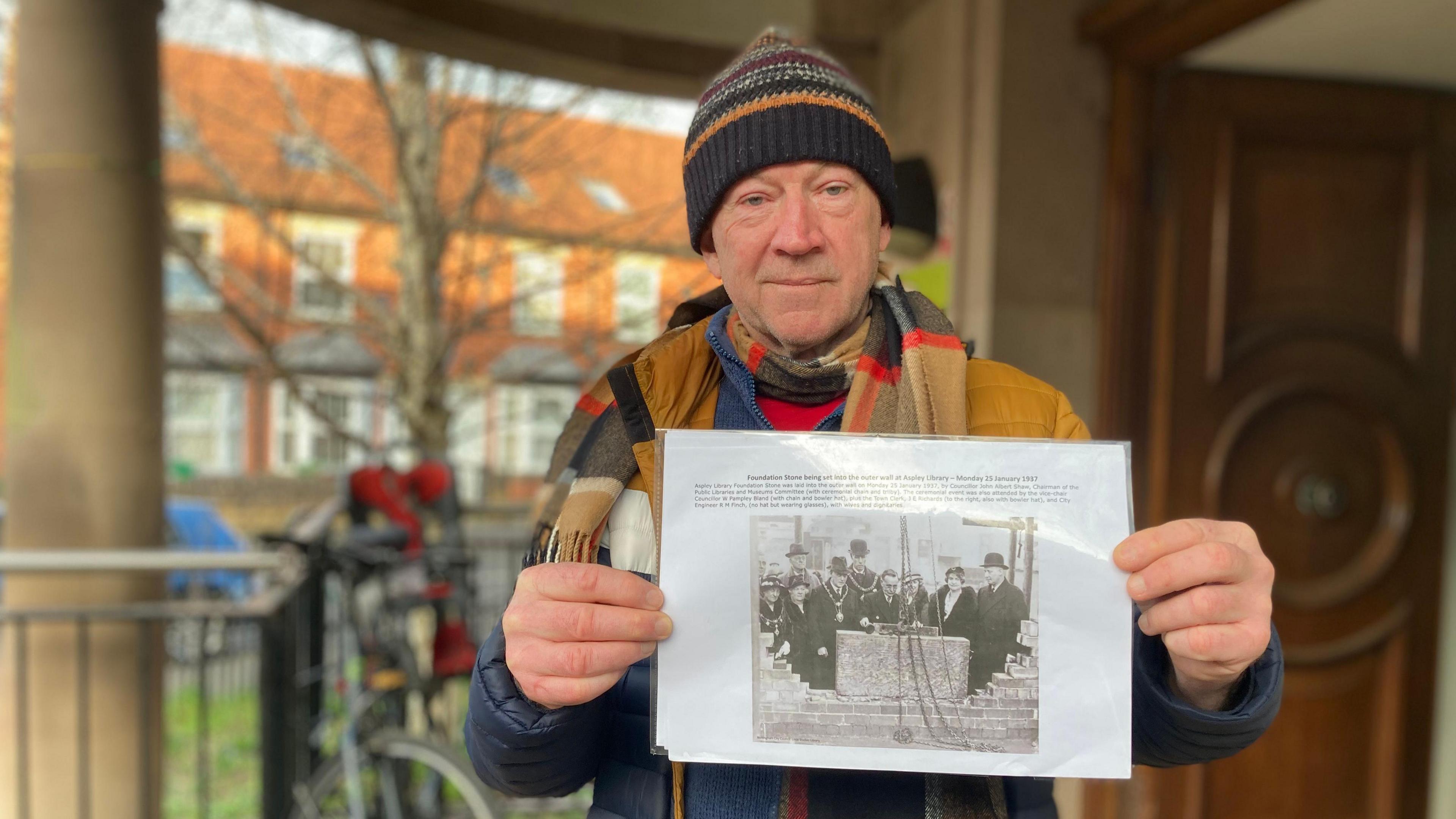 Campaigner Des Conway holding up a photograph of the grand opening of The Meadows library while stood outside it.