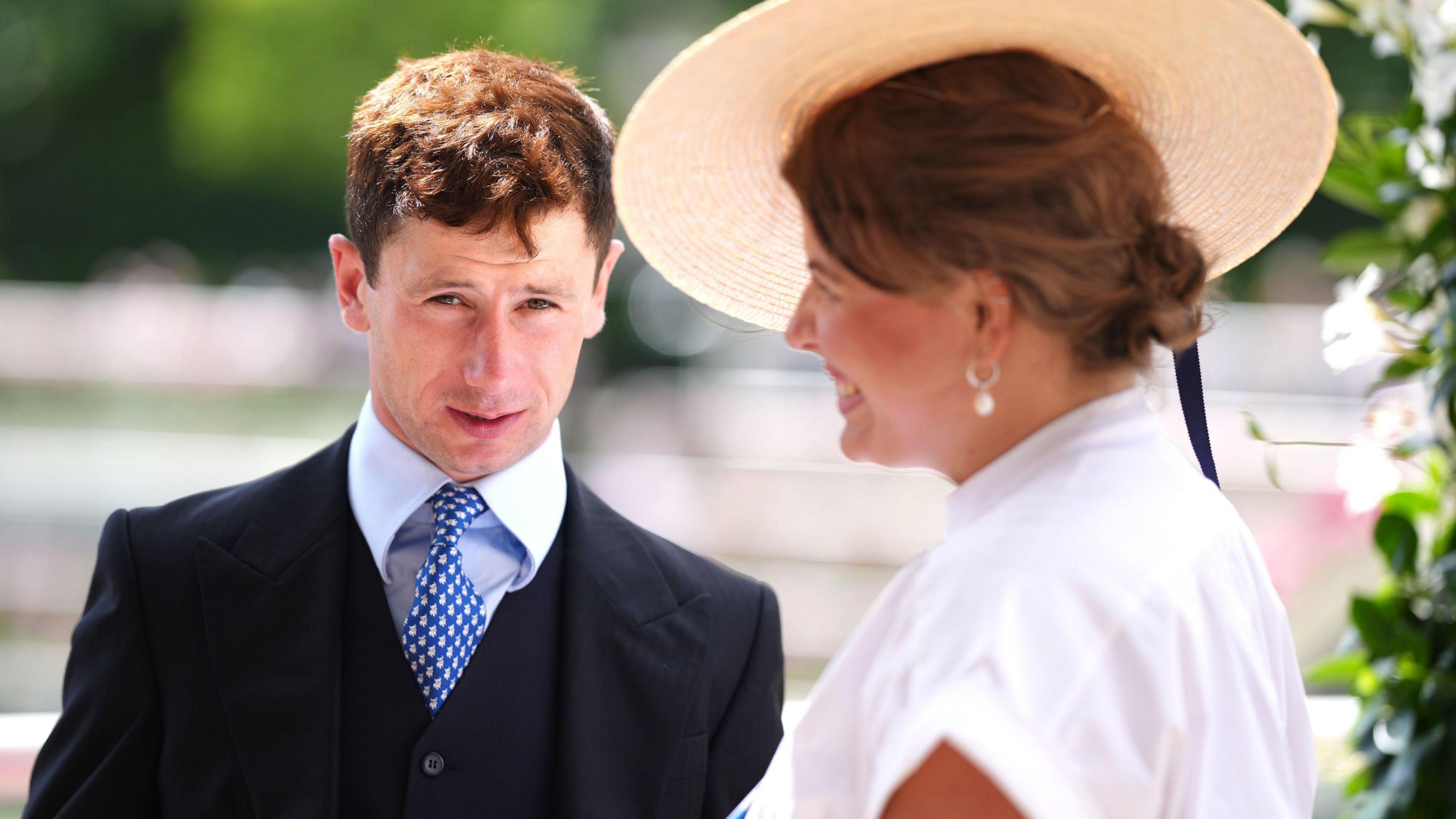 Oisin Murphy is looking towards the camera. He has short brown hair and wears a dark navy blue blazer and waistcoat, with a pale blue shirt and blue and white patterned tie. To his right a woman with brown hair tied in a low bun, wearing a white short sleeved shirt dress and white brimmed straw fascinator smiles and looks away from the camera.