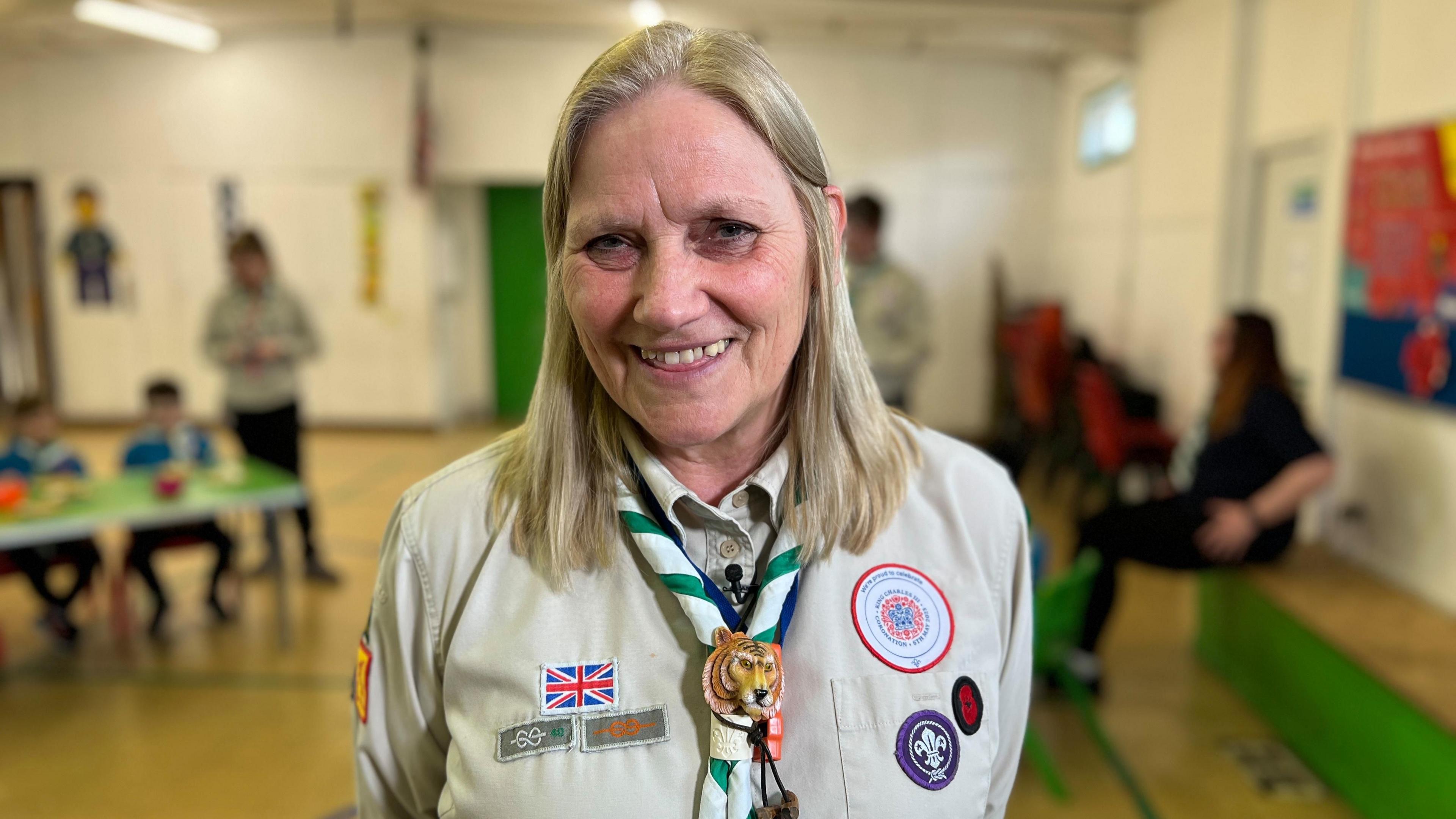 Nicky Taylor is smiling and looking at the camera. She has shoulder length blond hair and is wearing a scout uniform adorned with various badges.