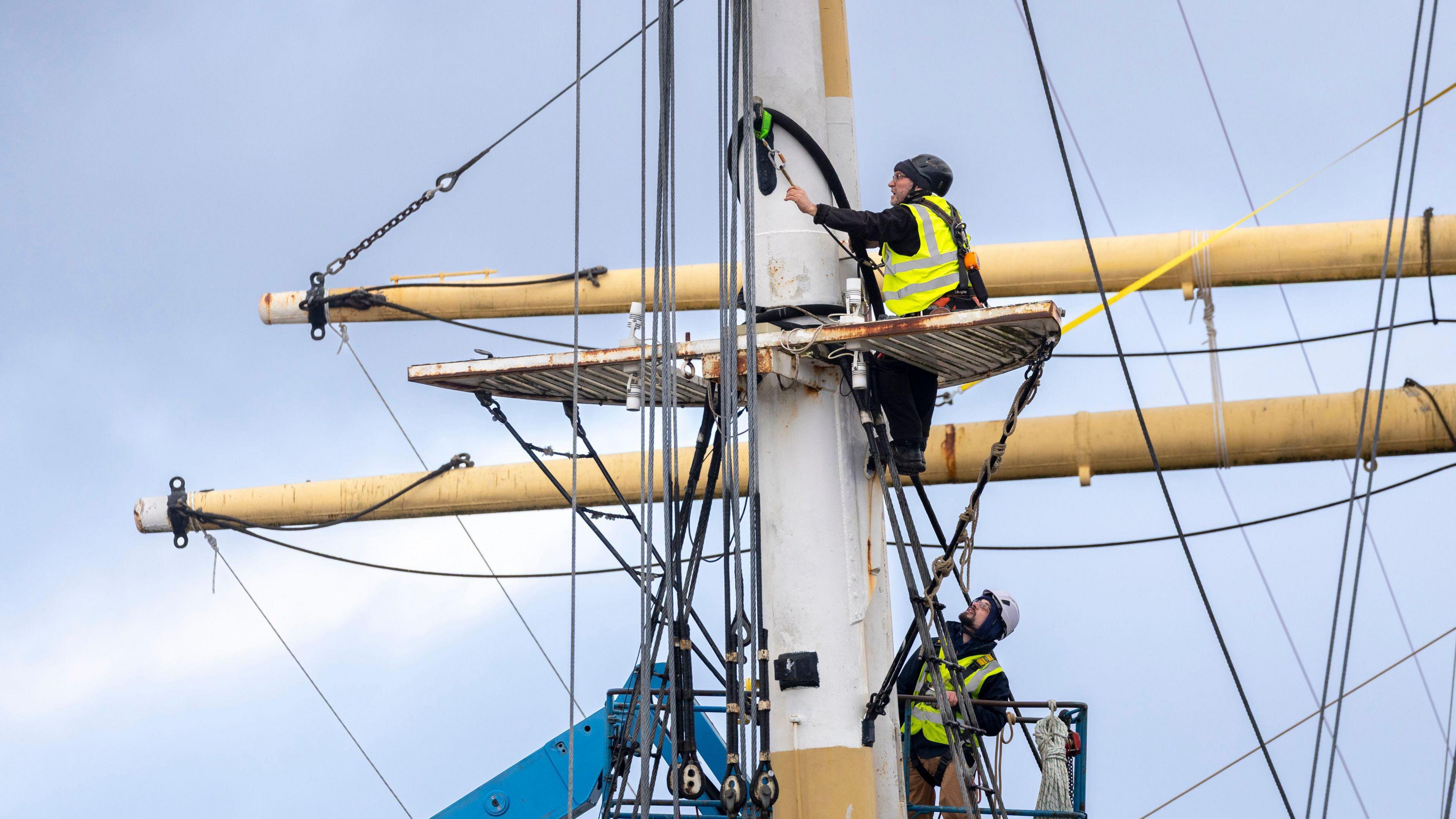 Two men in high vis yellow jackets, working on the mast of a large boat. They are helping to remove the masts for conservation work.