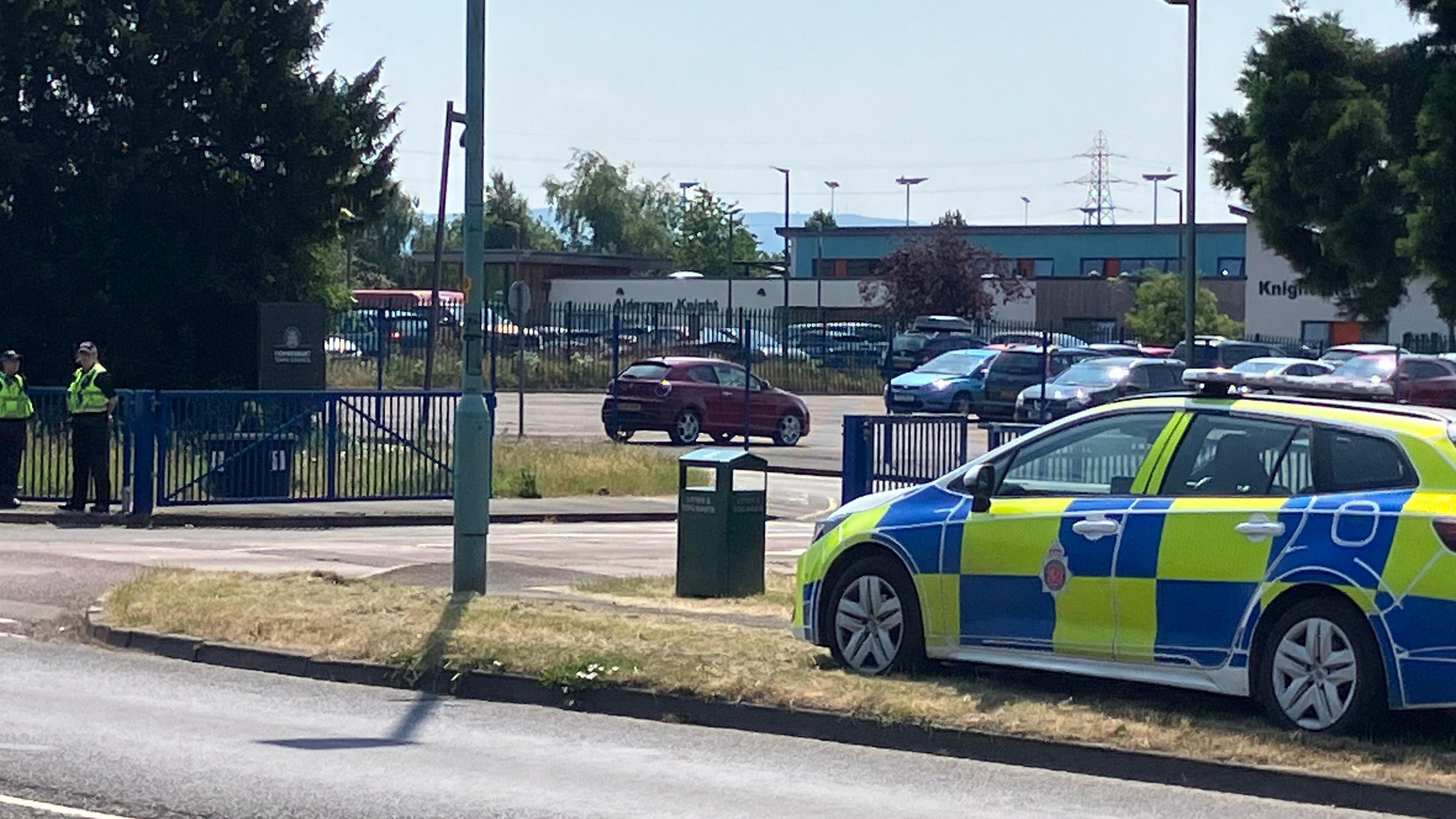 Entrance at Alderman Knight School in Ashchurch, Gloucestershire. A police car and two officers can be seen by the gates, and the school car park and building in the distance.