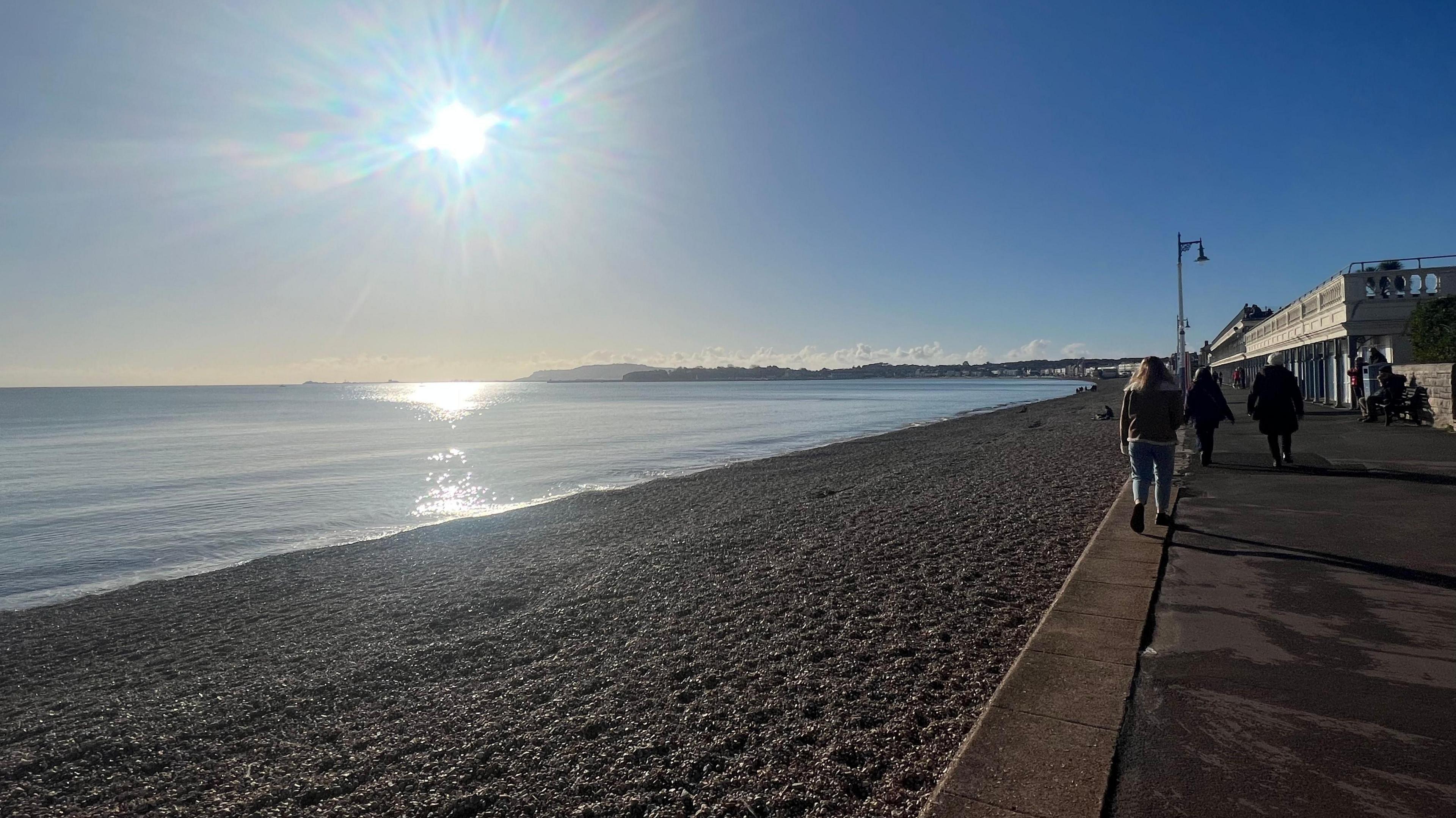 blue sea with stoney beach next to a concrete esplanade. People are walking alongside the beach. A low sun shines in a cloudless sky.