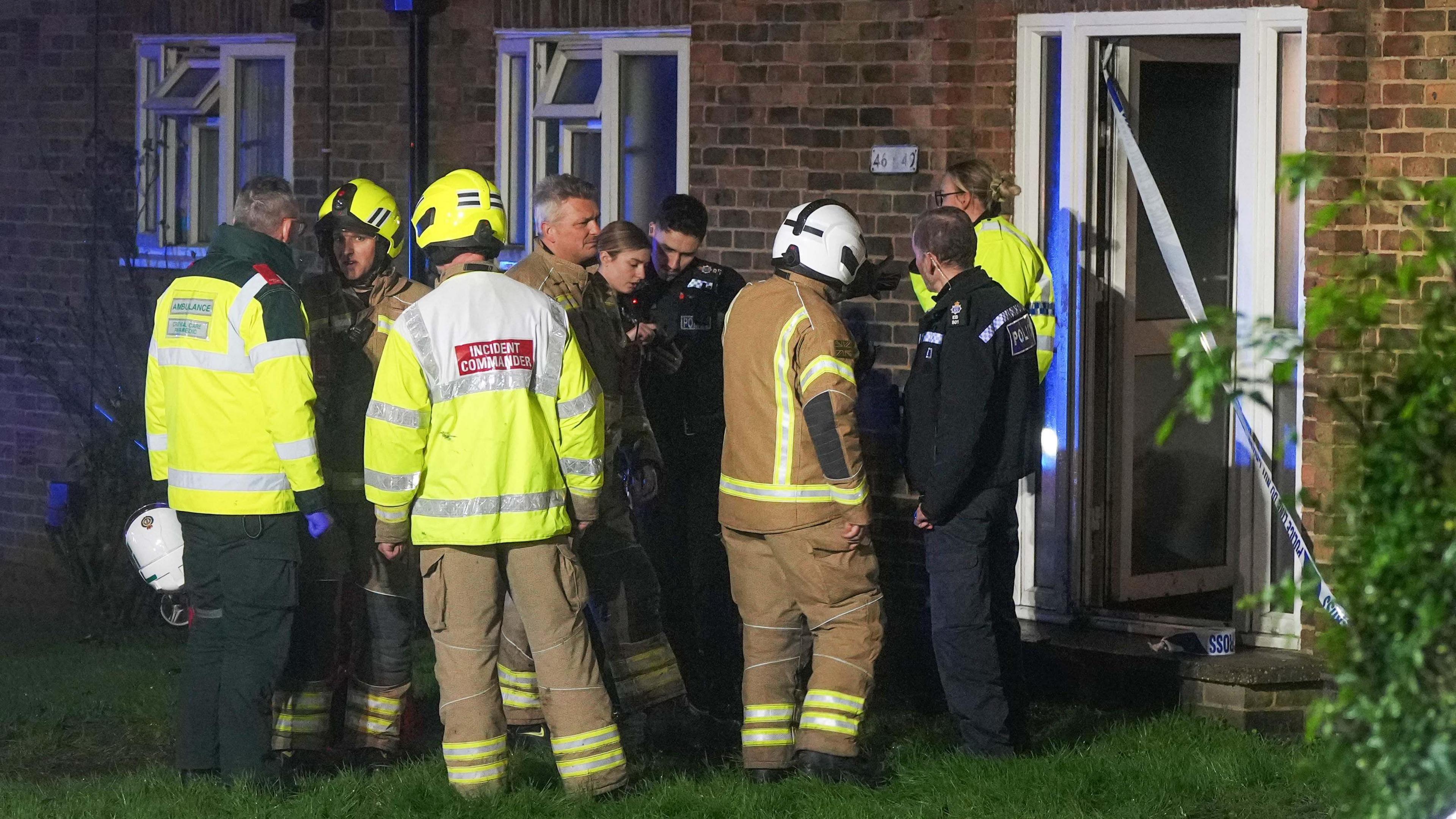 A group of fire fighters and police officers gather outside the door of a block of flats that has caught fire in Steyning. The front door is open but sealed off by police tape.