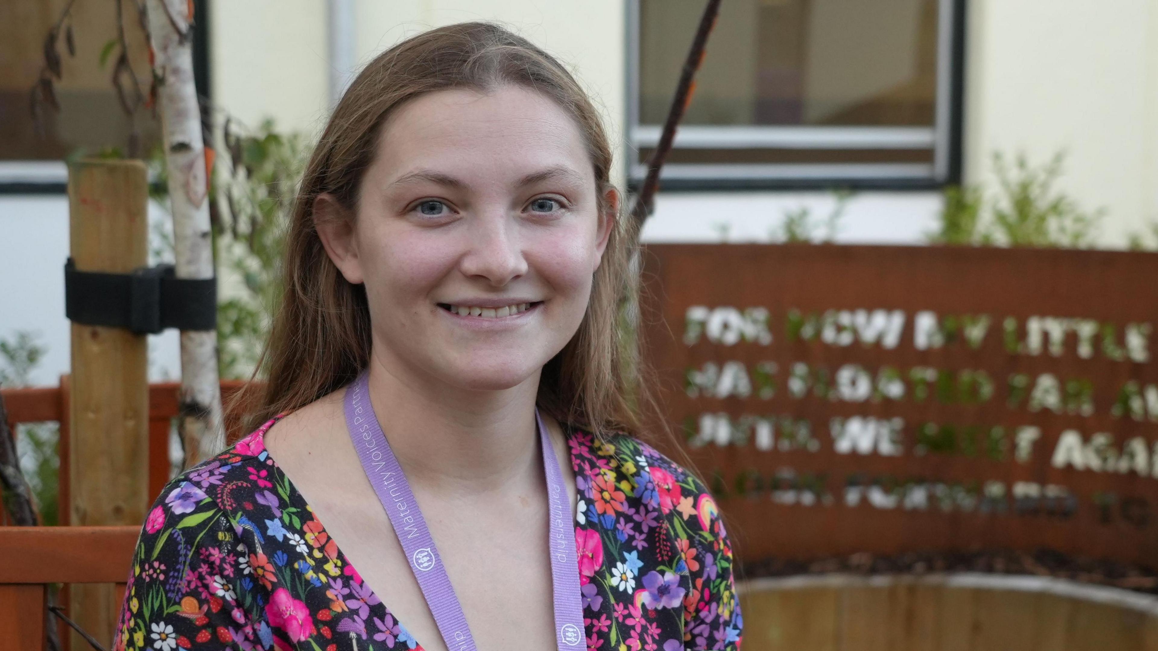 Emily Lunny wearing a black dress with pink, purple, orange, yellow, blue, red and green flowers on it. She has long brown hair and is wearing a purple lanyard round her neck. She is sat outside in the garden.