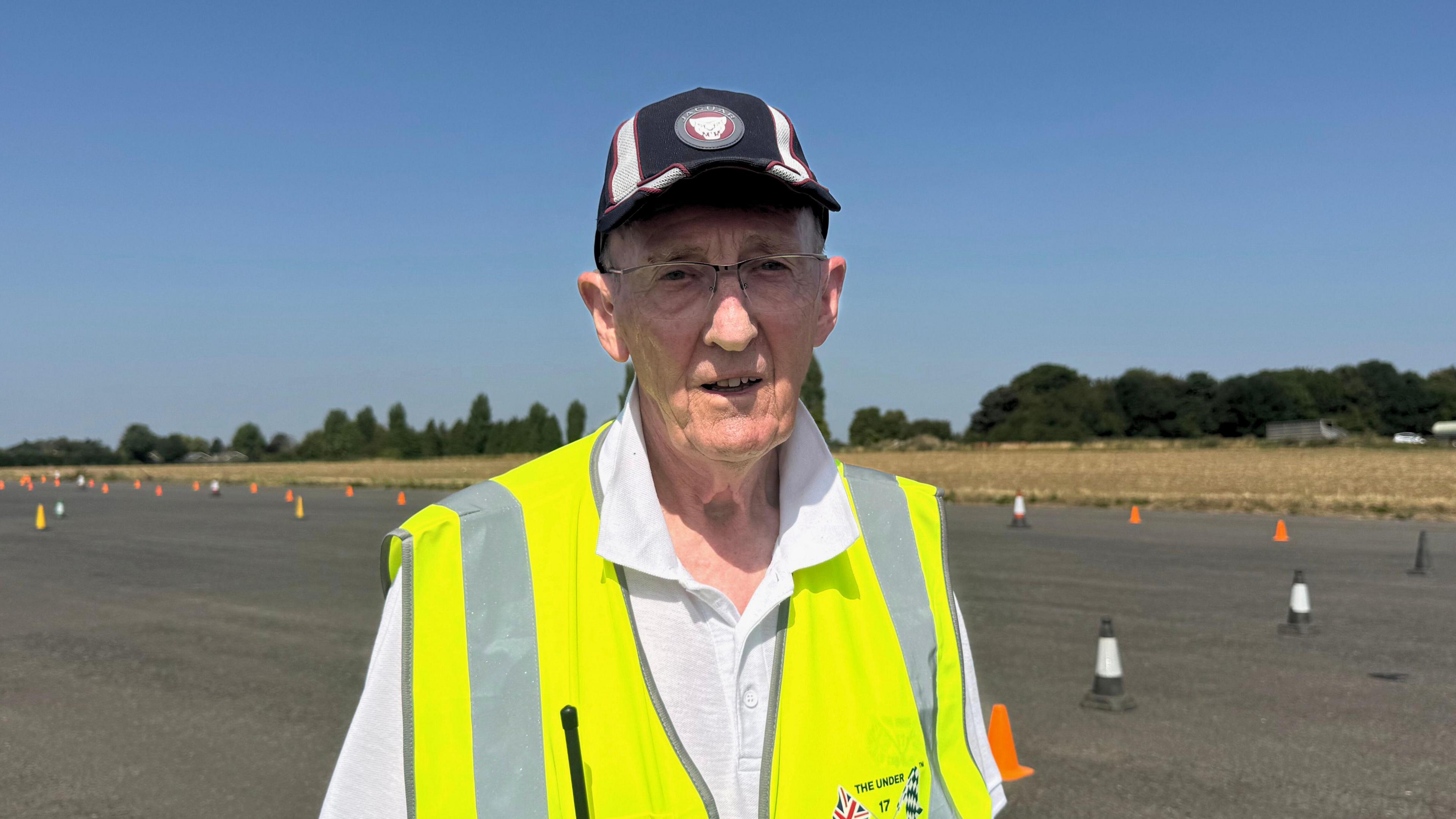 A man in a hi-vis vest stands on tarmac at an airfield. The tarmac is lined with orange cones.