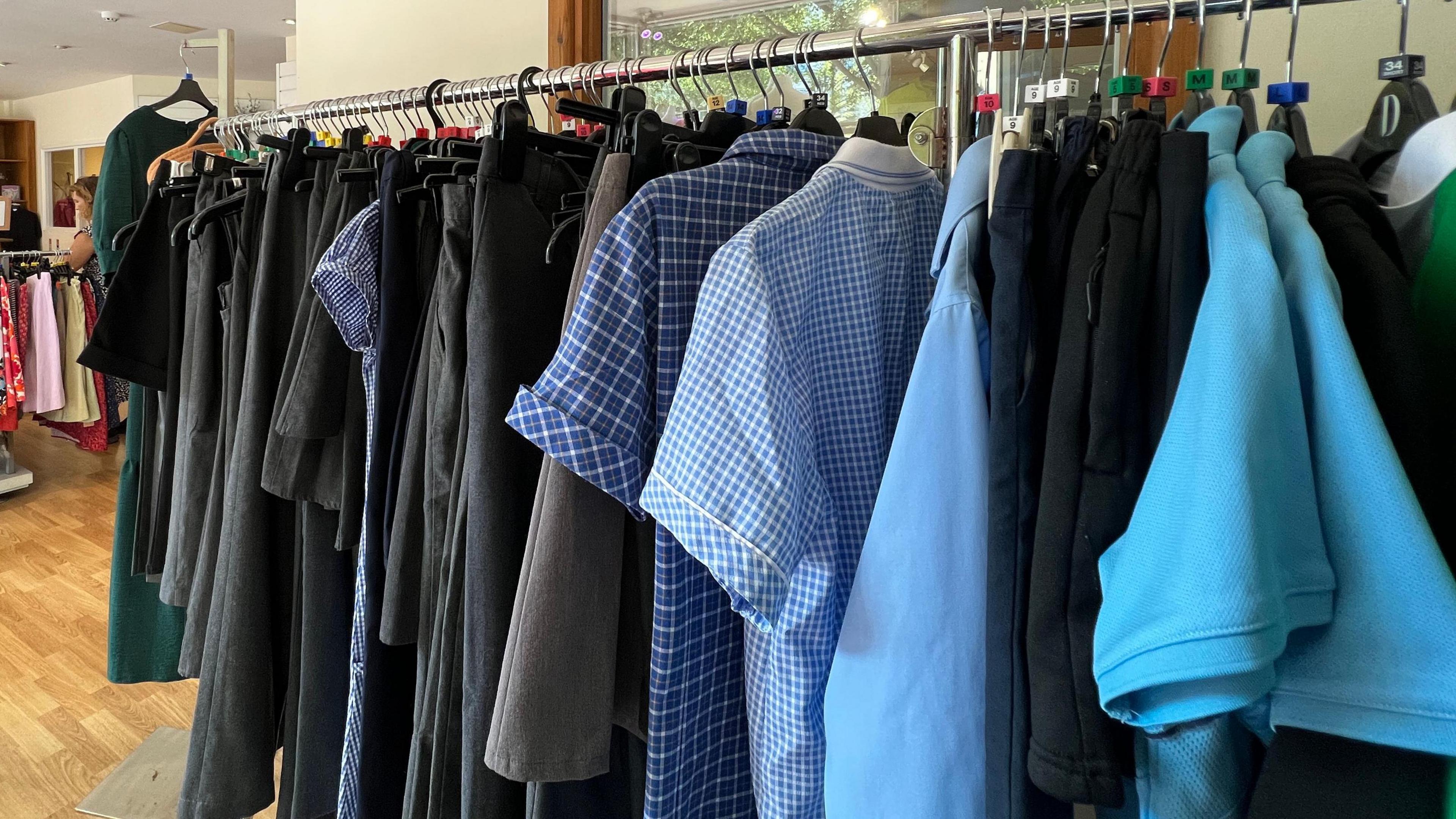 A clothing rack in an Autism Jersey shop has a range of grey trousers and skirts, blue and white checked, short-sleeved shirts and dresses, pale blue short-sleeved tops and black trousers and skirts.