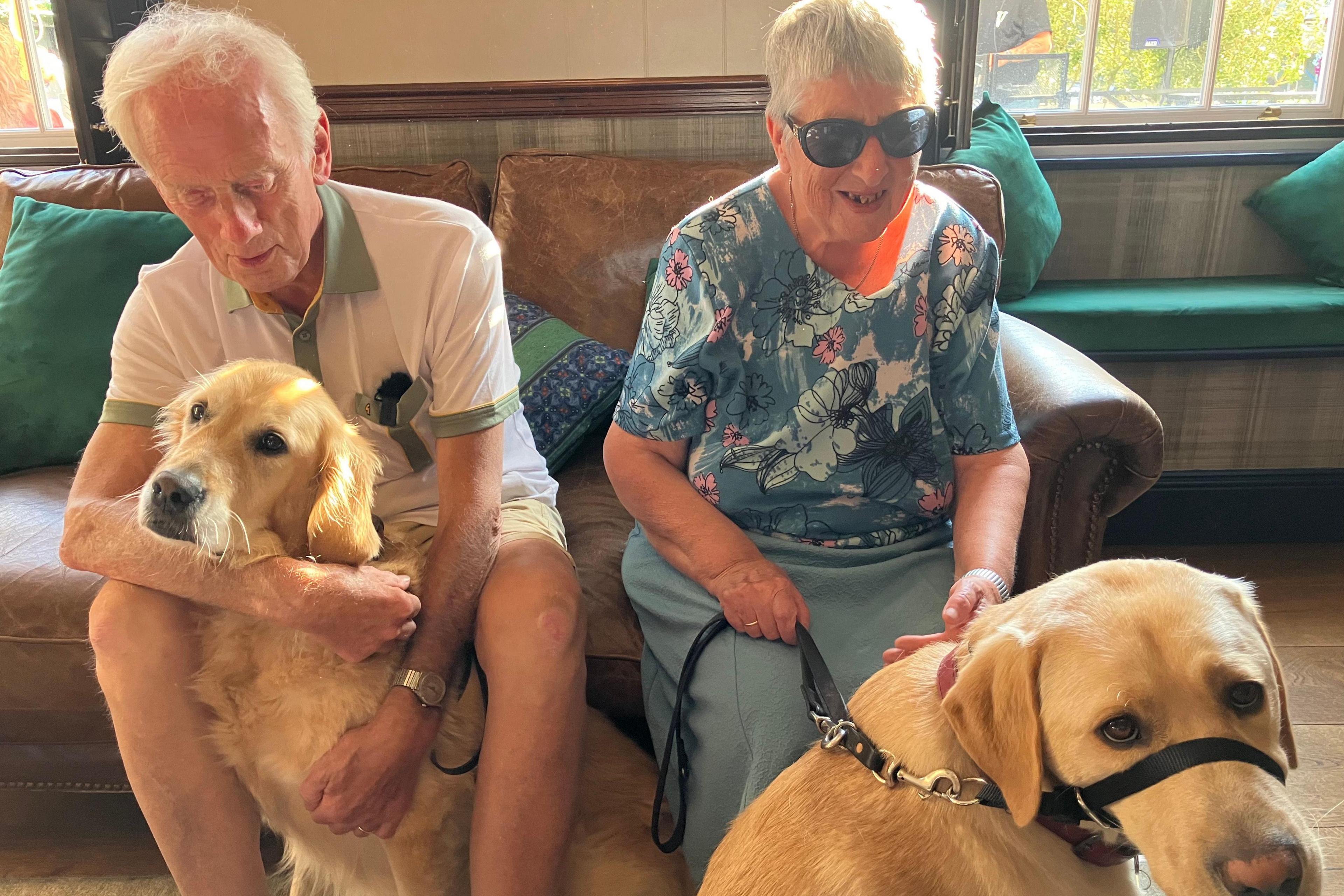 Jeff and Janie Johnson feature with their guide dogs. Jeff wears a white T-shirt and shorts, hugging his golden retriever guide dog, Lilly. Janie, dressed in a blue and pink skirt and blouse, wearing dark glasses, strokes Davy, her golden Labrador.