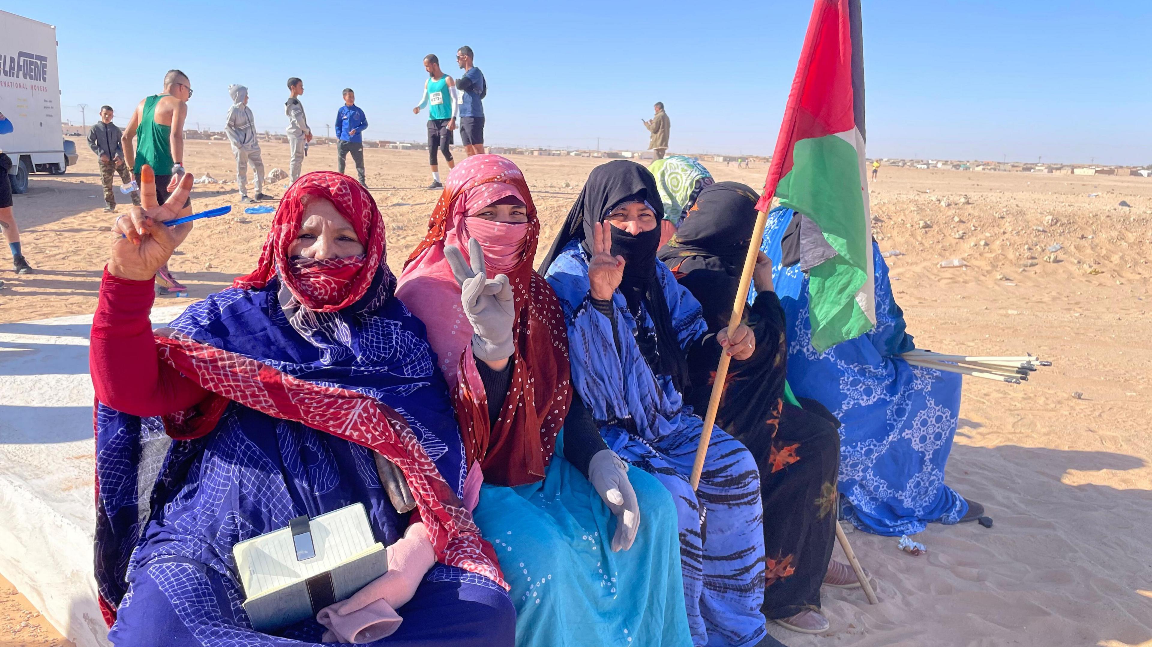 Women, in red head scarves and blue dresses, hold up their hands at a refugee camp in Algeria