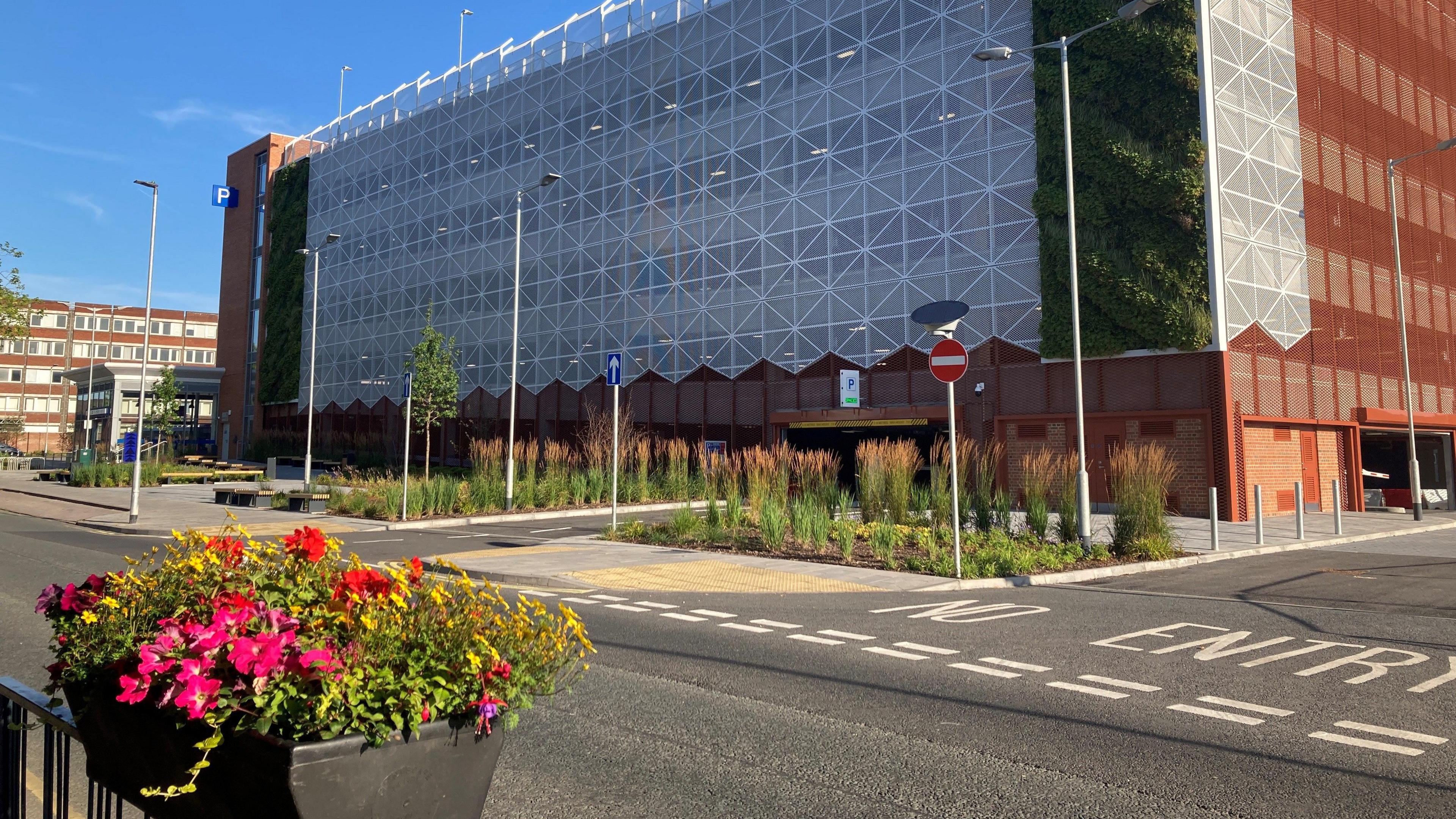 A view of the multi-storey car park in Crewe. It is a large sqaure block with white criss-cross panelling on one side and red around the rest of the building. On the far side, you can see a block stairwell with a large P on the top of the section. There are roads around the car park and a building that looks like flats behind it.