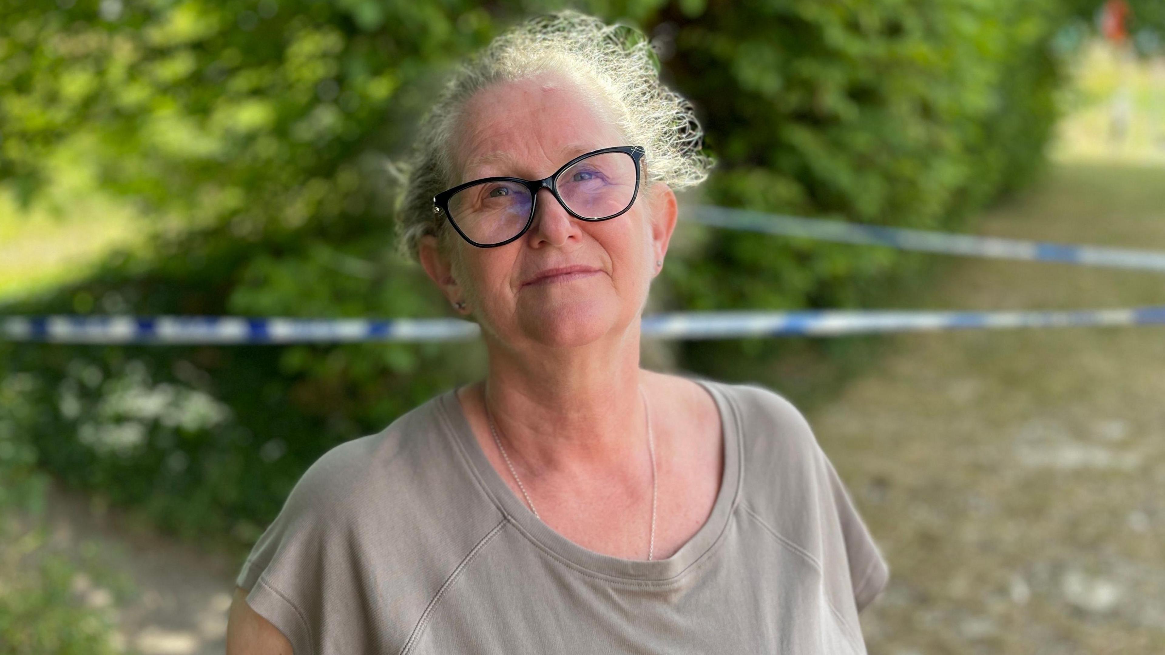 A woman wearing glasses and a grey top is stood in front of some bushes. Between her and the bush is blue and white police tape.