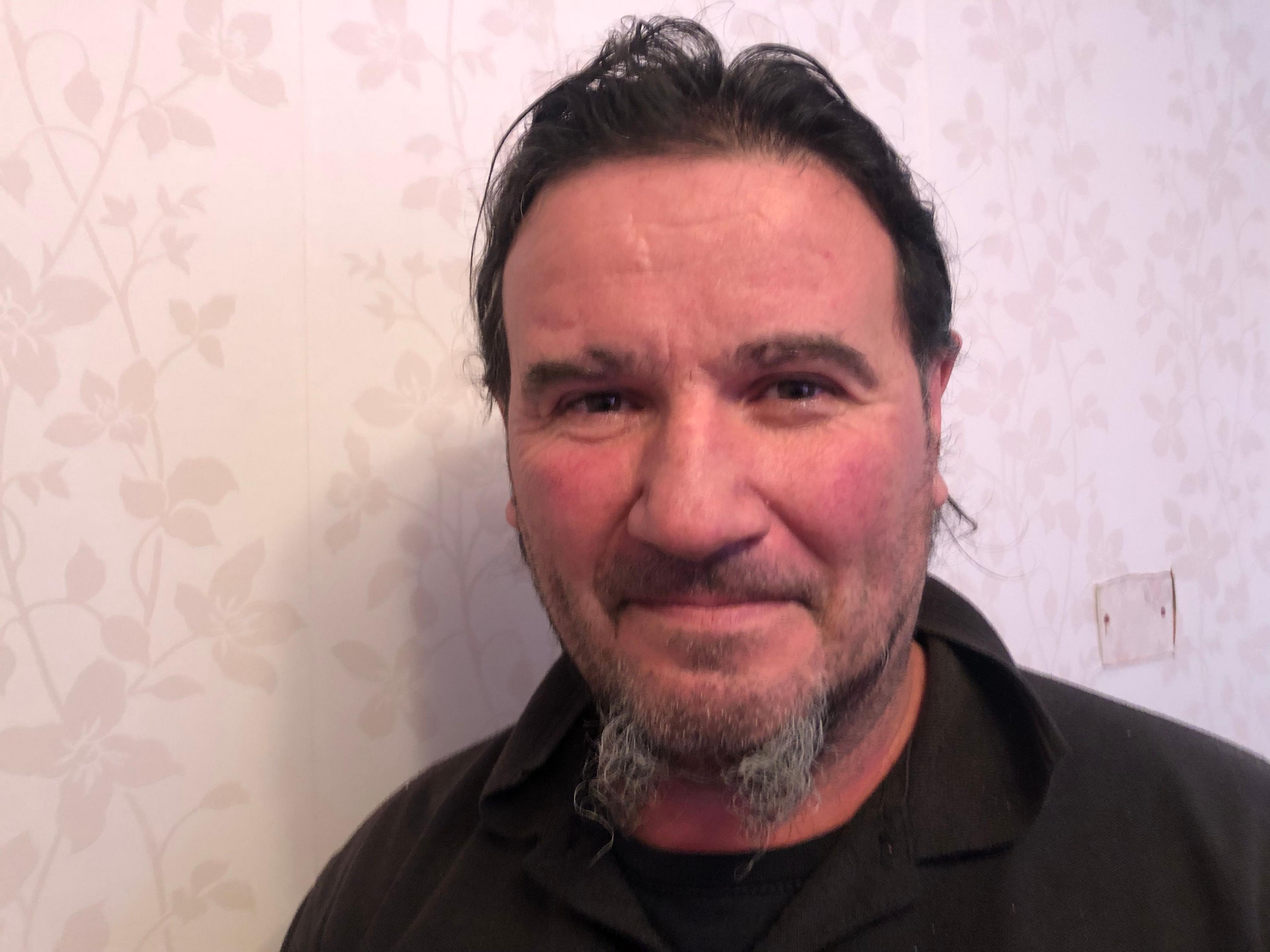 A head-and-shoulders picture of Richard Baxter. He has a wispy black and grey beard and moustache, his dark hair is swept back. He is wearing a dark jumper with black shirt underneath. He is standing in a room with light plain floral wallpaper.