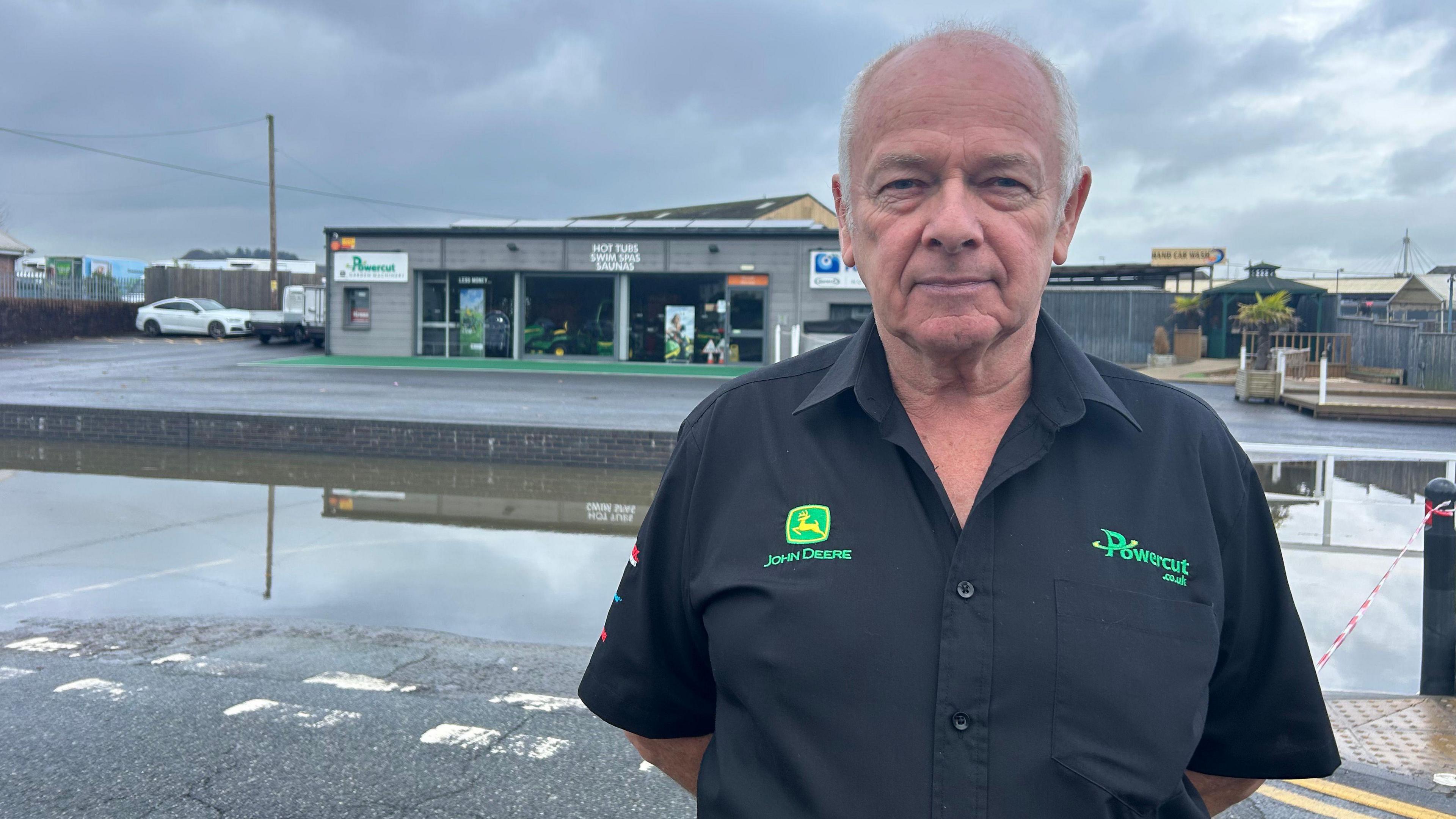 A man with short grey hair, wearing a black top with green writing on it standing in front of his shop and a flooded street