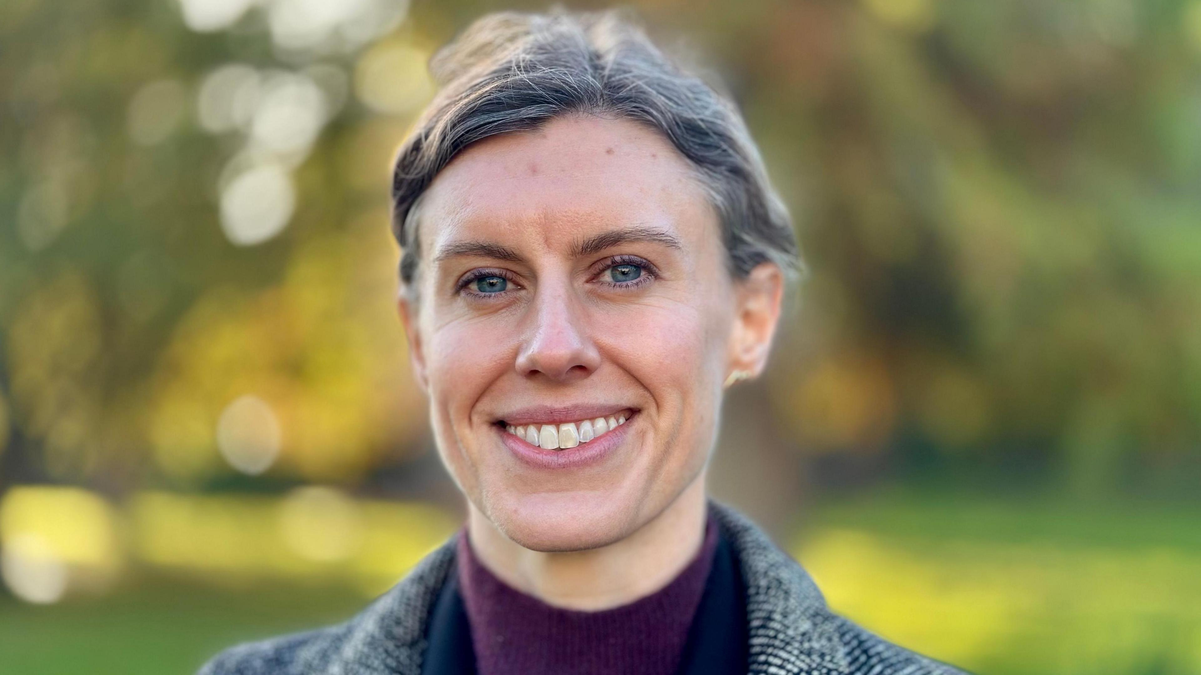 A woman approximately in her late thirties with pulled back brown hair, flecked with grey wearing a polo neck jumper and winter coat pictured in a park