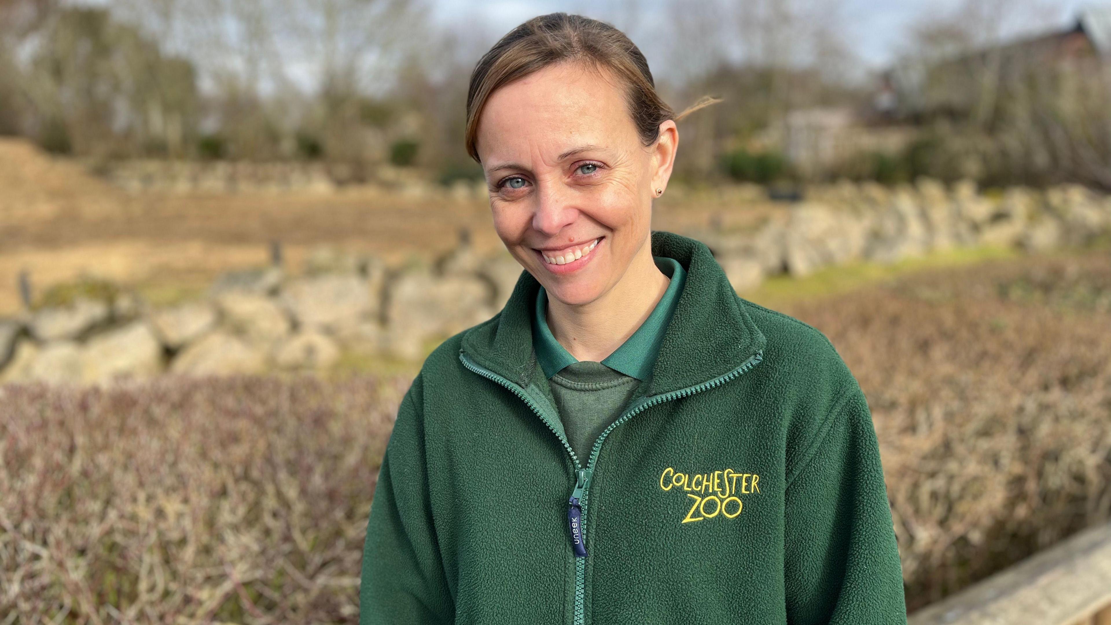 Rebecca Moore, zoological director of Colchester Zoo, looking at the camera and smiling. She is wearing a green polo shirt, jumper and fleece jacket with yellow Colchester Zoo branding on her left chest. She is standing in front of an outdoor animal enclosure.