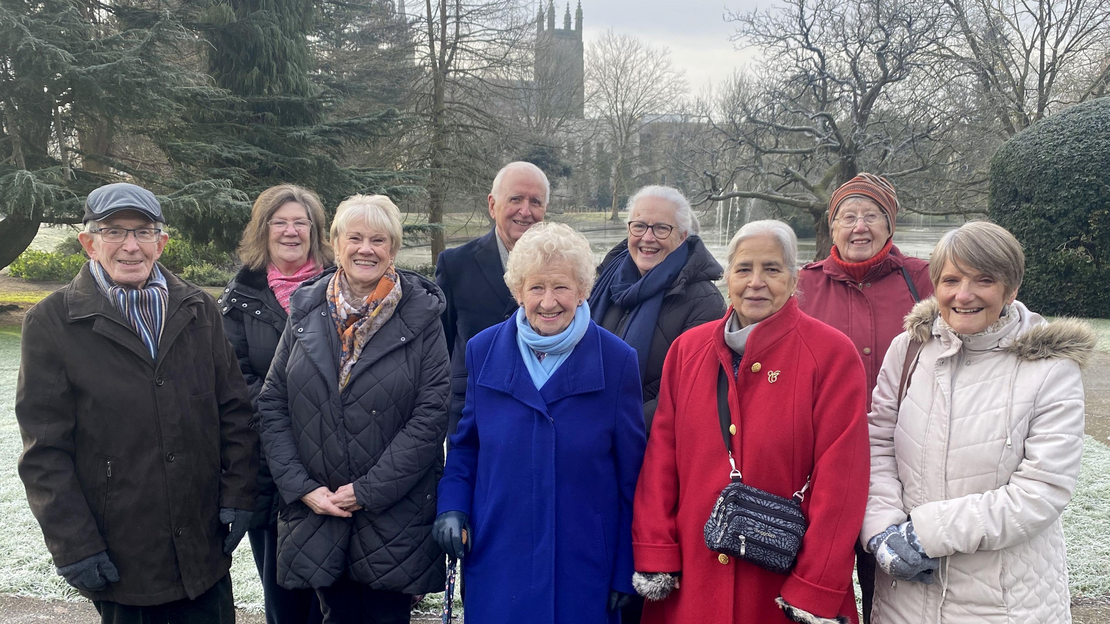 Nine people in a park. Seven women and two men. They are all people who contributed to the memories of the lights festival project. They are all wearing coats and scarves and the ground behind is frozen.