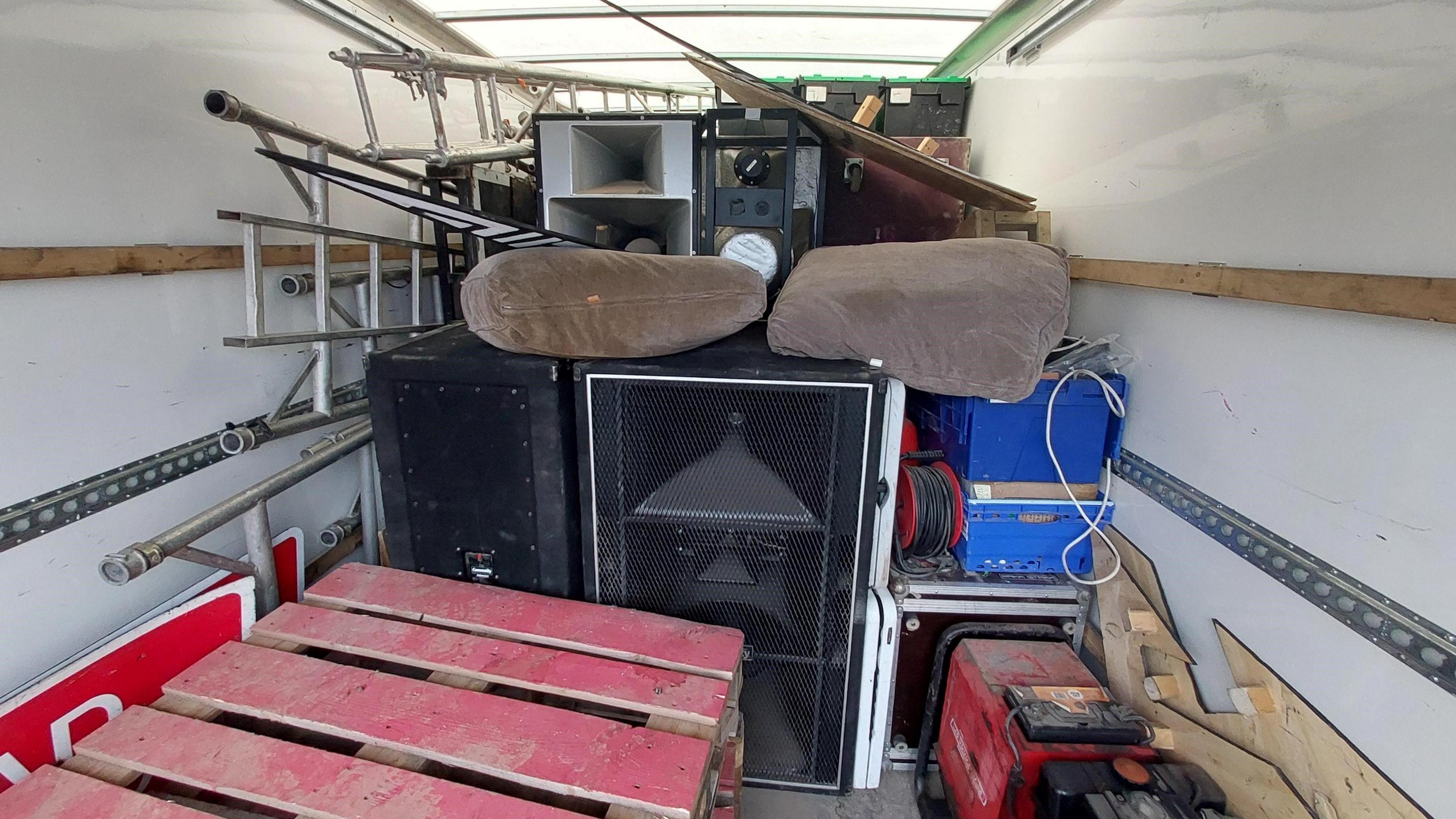 The inside of a  white van with lots of clutter including a table, poles, cushions and large speakers. The objects are piled on top of each other. 