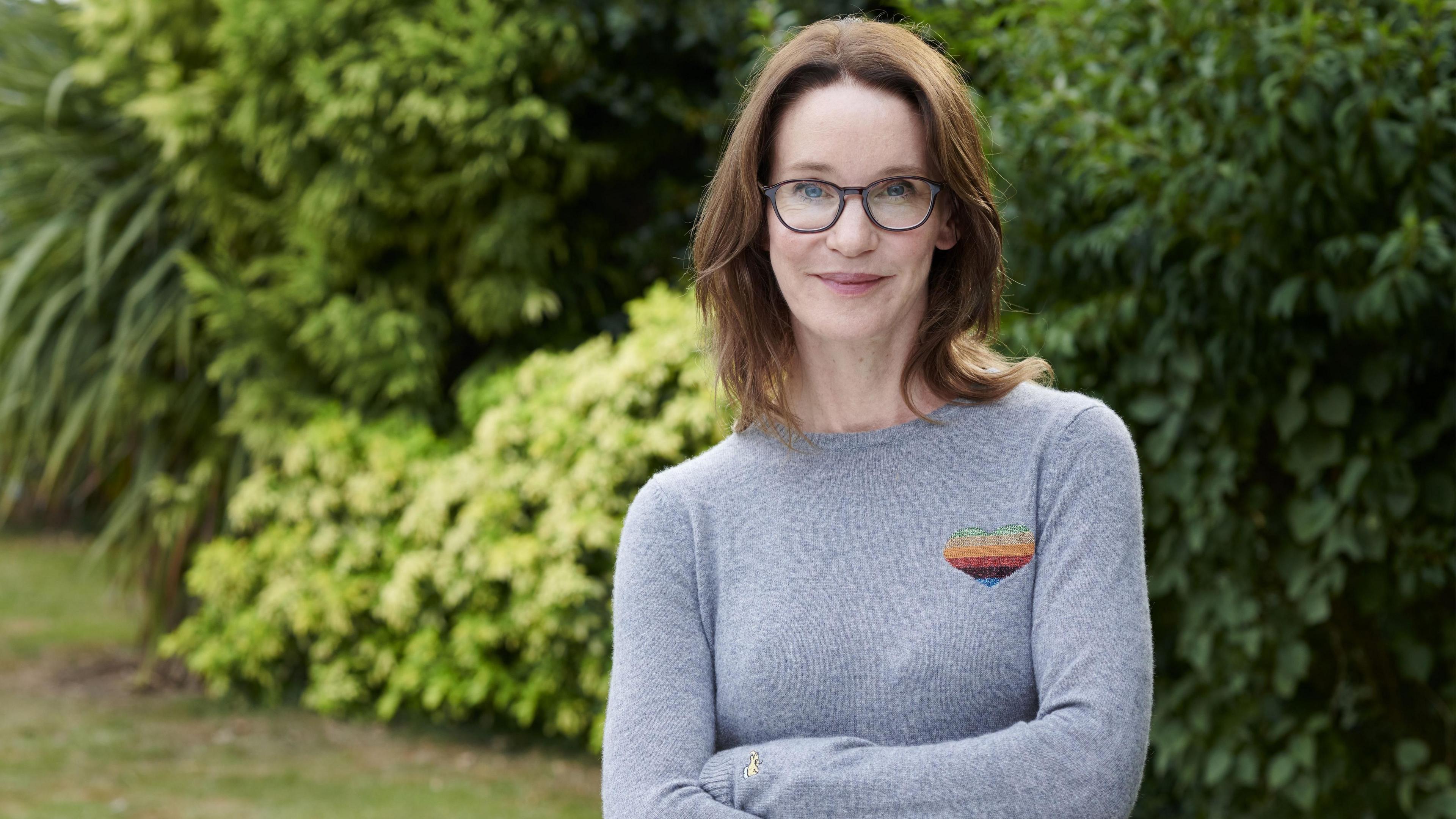 A woman with shoulder-length brown hair and glasses, wearing a light blue/grey jumper stands with her arms folded in front of trees and bushes, smiling at the camera.