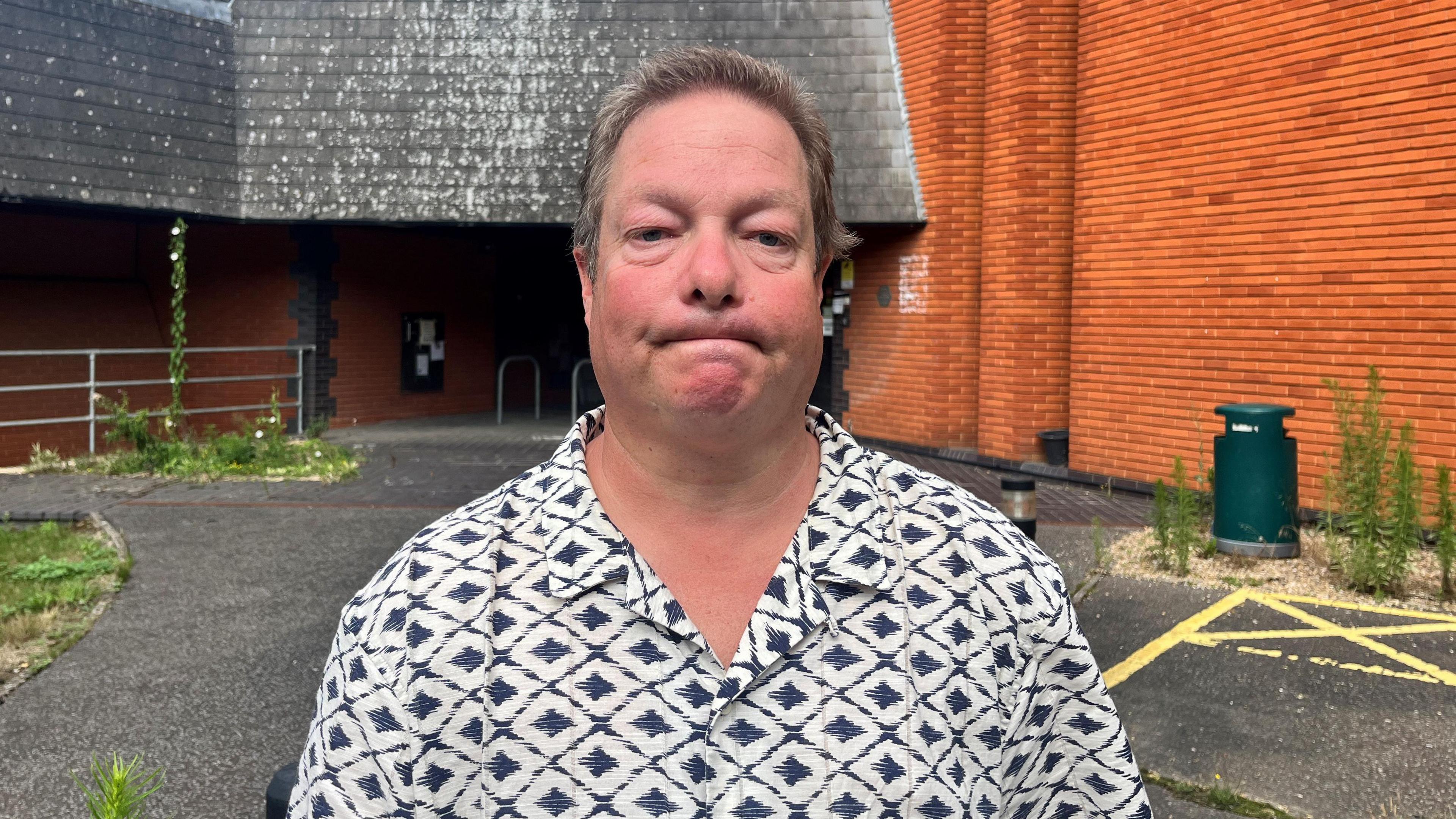 Paul Rochester is standing in front of South Norfolk House. He's wearing a mostly white shirt with dark diamonds on it. Behind him, we can see that parts of the ground outside the building have become overgrown.