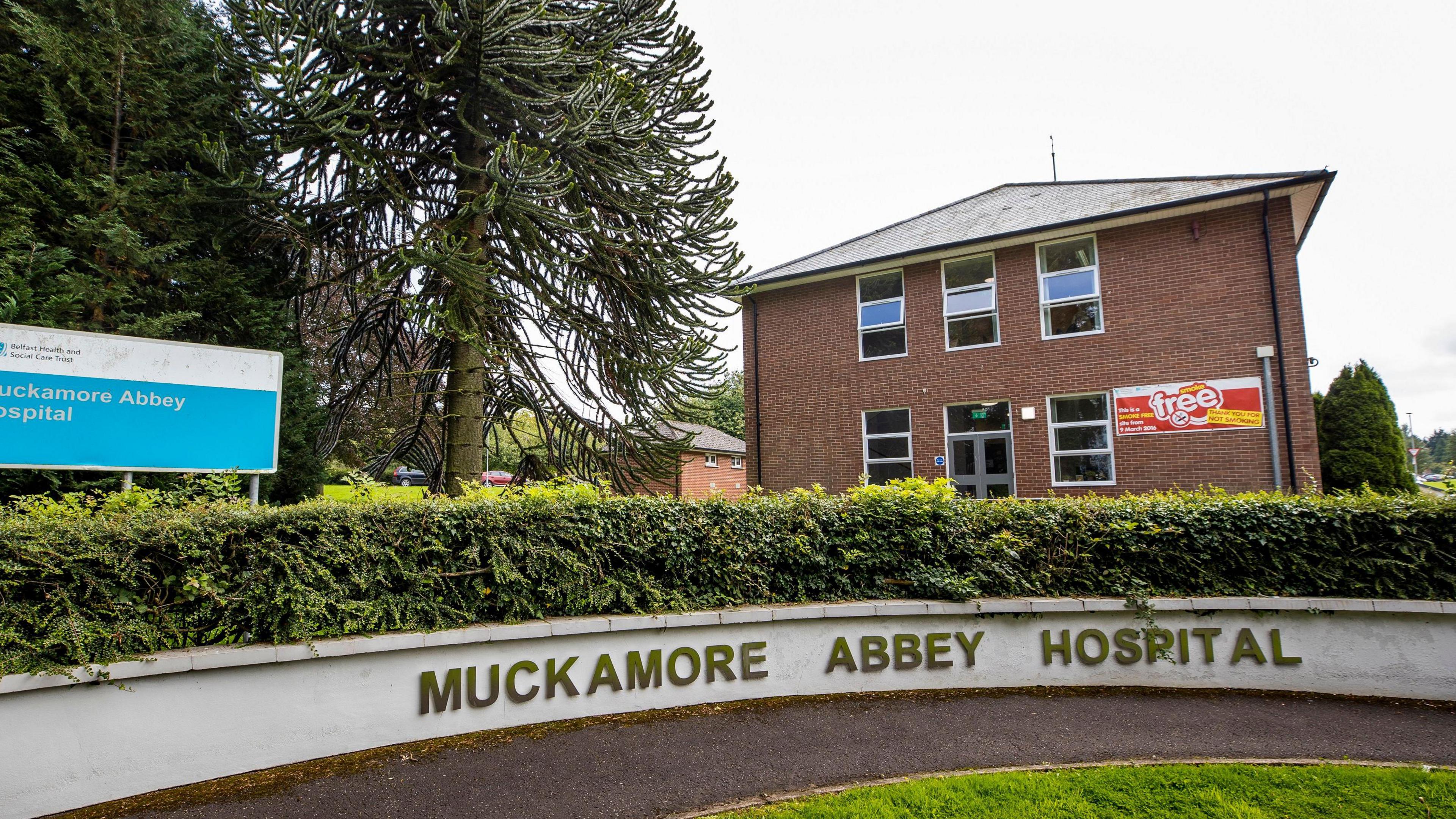 In the foreground is a curved white wall which has the words Muckamore Abbey Hospital out on it. Behind the wall is a hedge and trees and a  red-bricked building.