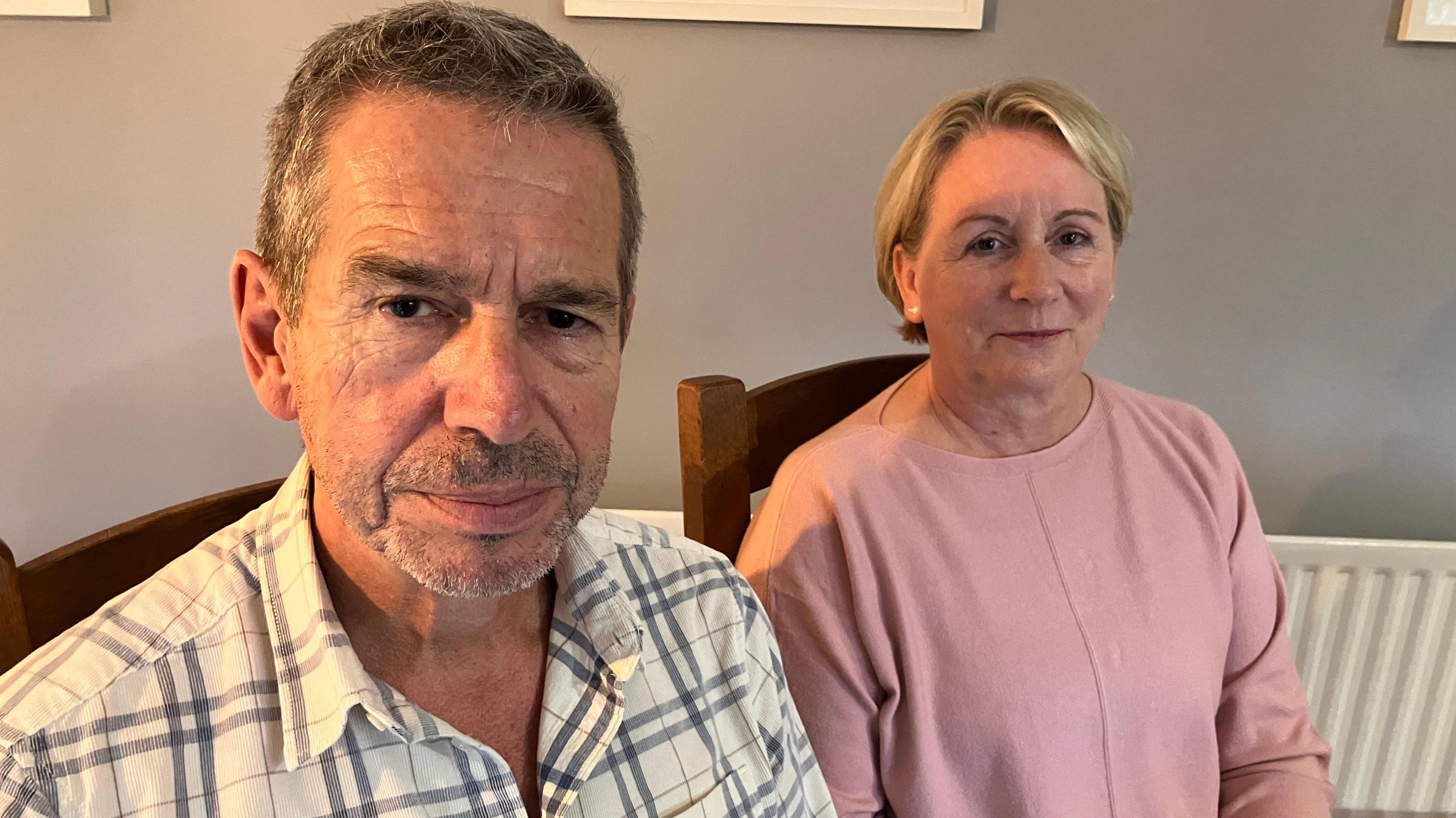 A man in a grey and white shirt and a woman in a pink top. Both are sat in wooden chairs and looking at the camera.