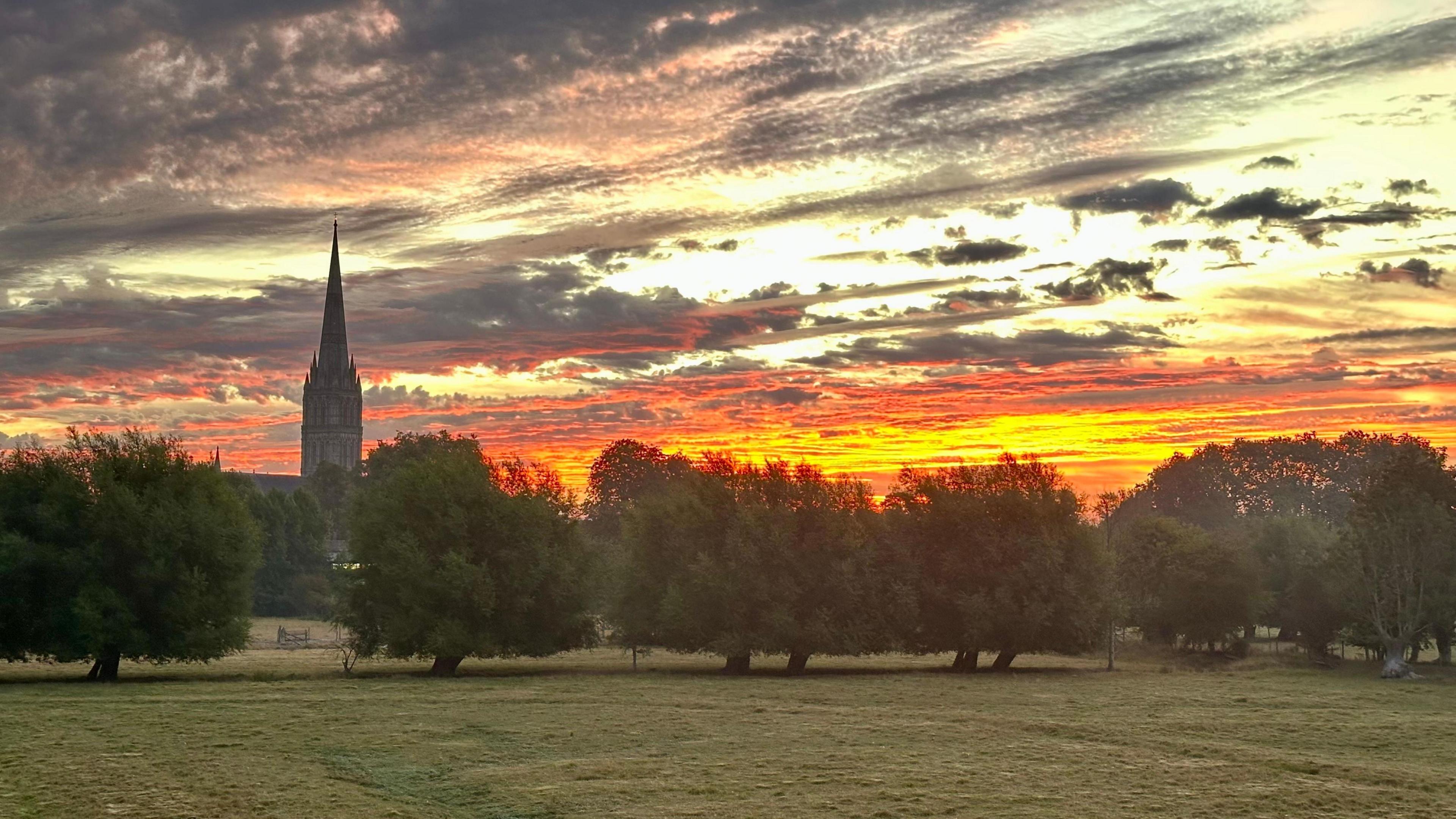 A dramatic sunrise with broken clouds delineating different hues of light in the morning sky, ranging from white to deep pink. Salisbury Cathedral and a line of trees can be seen in the background.