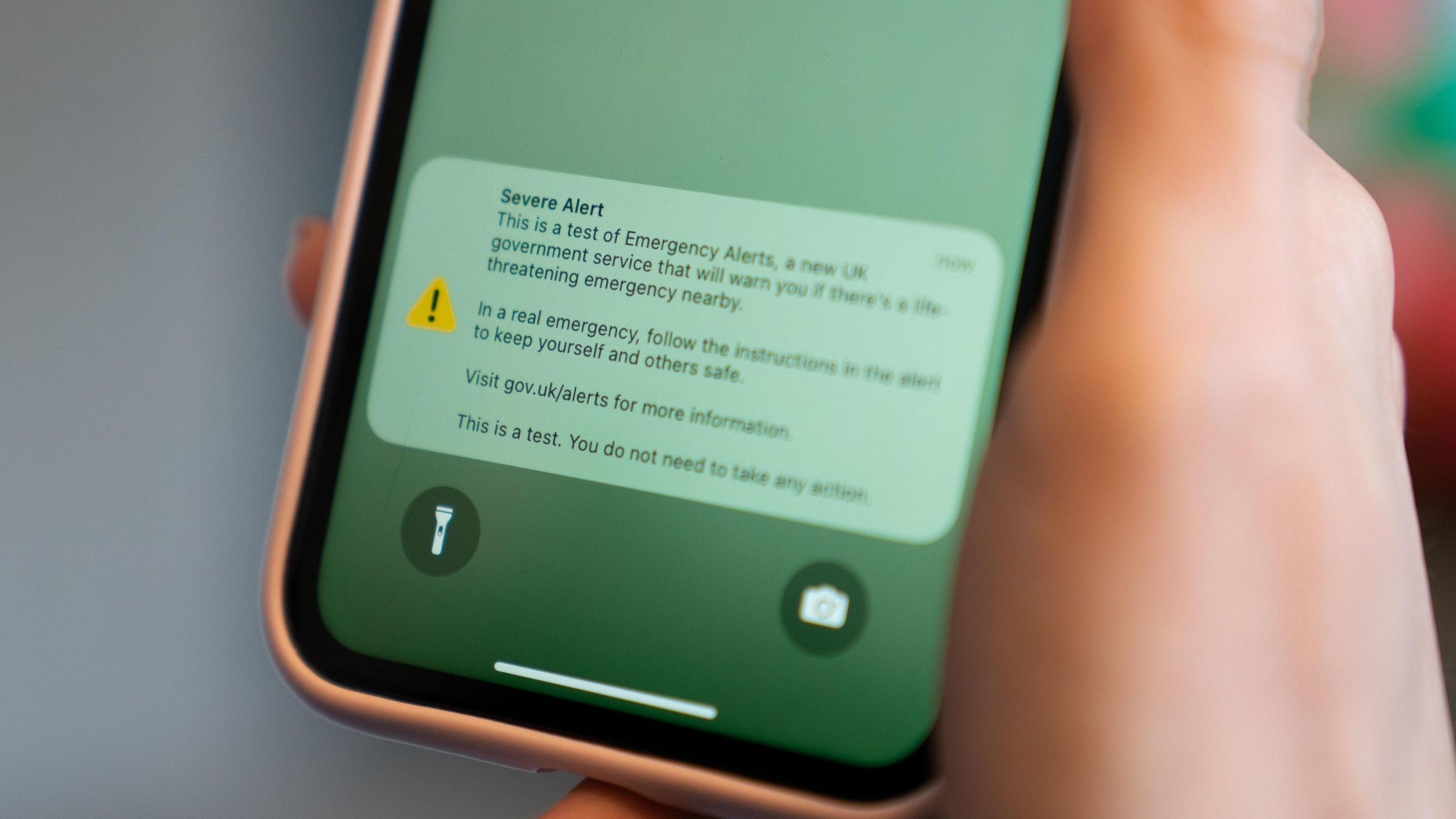 Image of a hand holding a phone. On the phone reads an emergency alert: "Severe alert. This is a test of Emergency Alerts, a new UK government service that will warn you if there's a life-threatening emergency nearby. In a real emergency, follow the instructions in the alert to keep yourself and others safe. Visit gov.uk/alerts for more information. This is a test. You do not need to take any action."